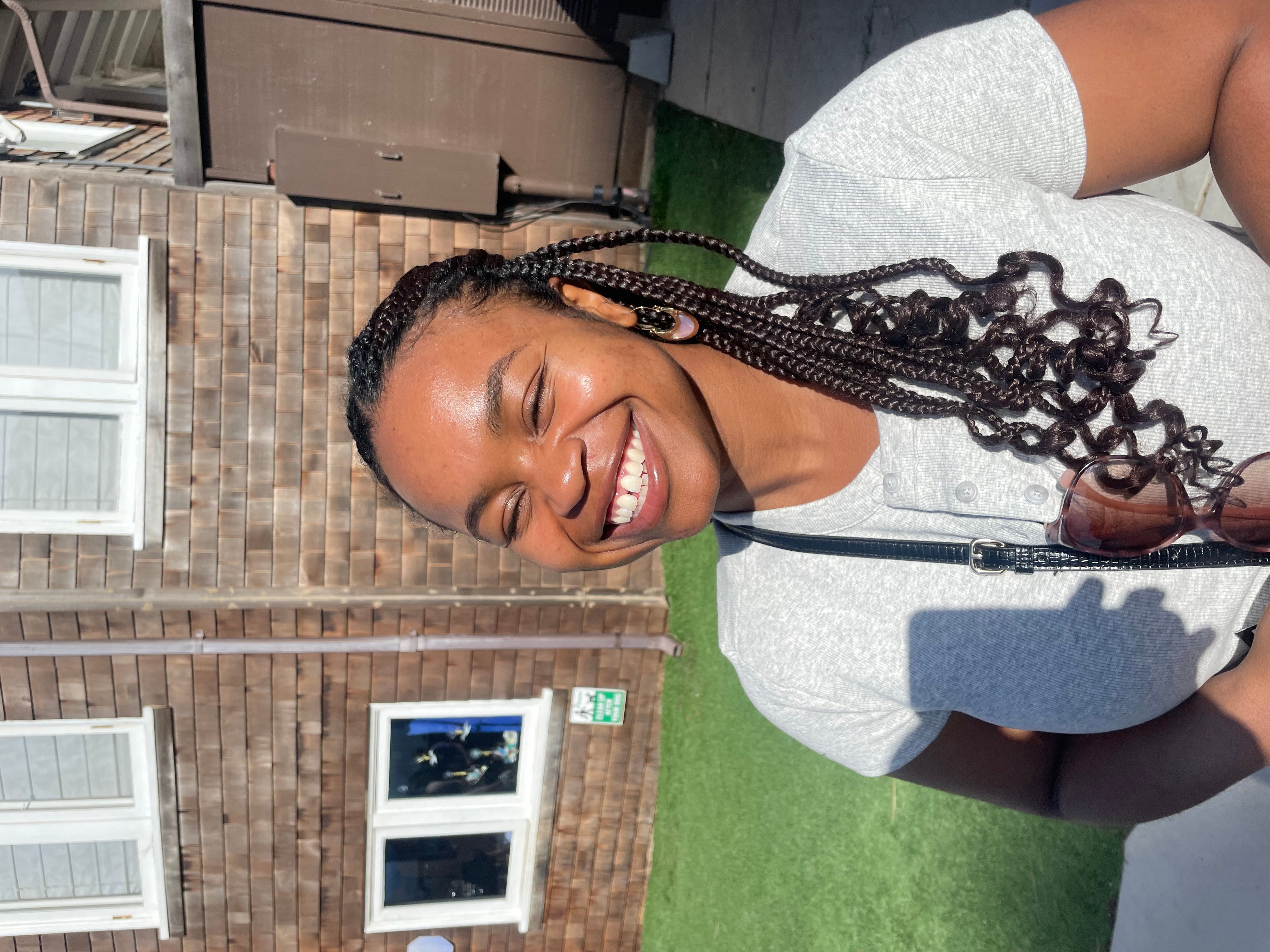 Smiling woman with braided hair outdoors, standing in front of a brick building with windows, wearing a light-colored top.