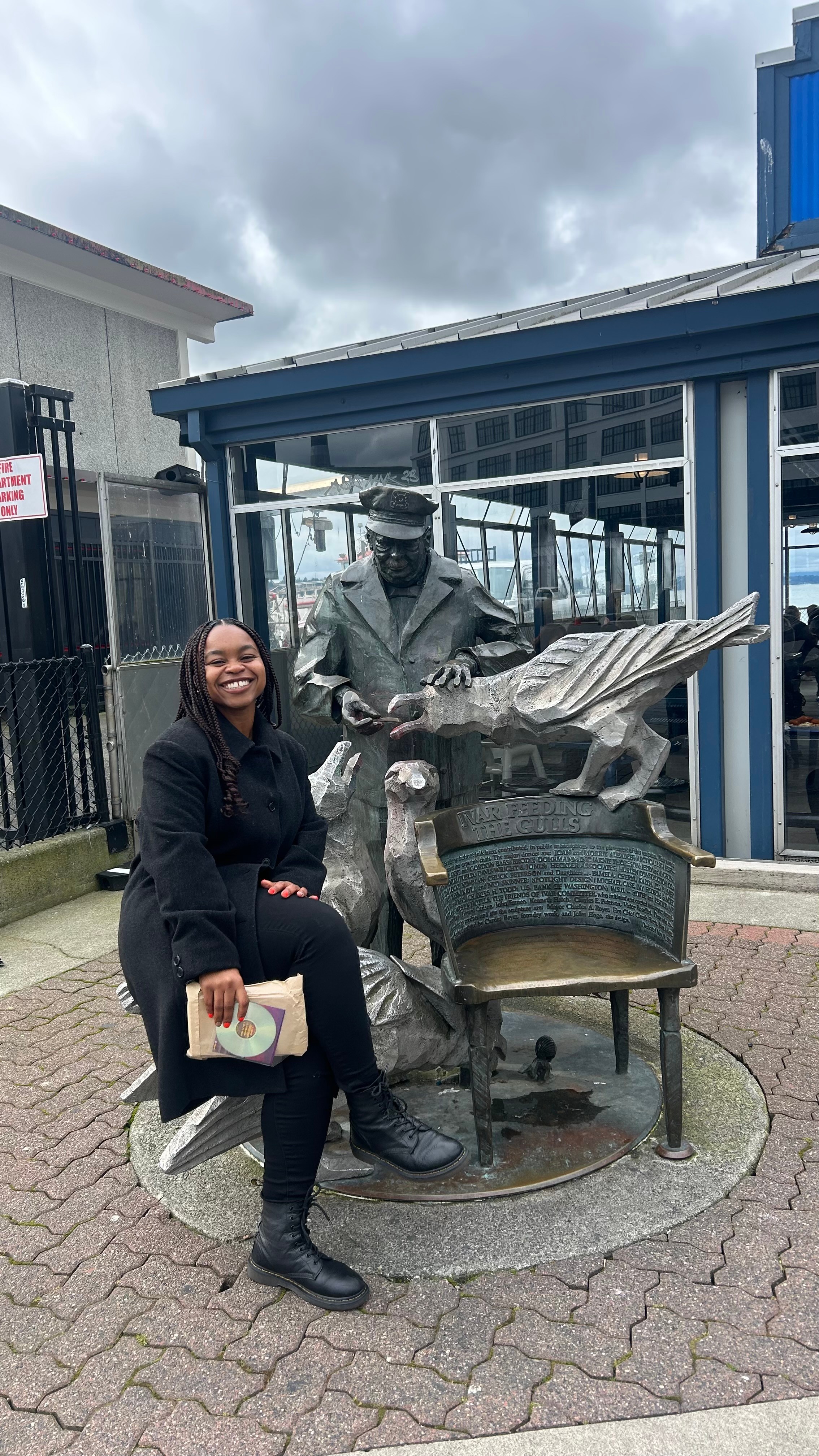 Woman sitting on a bench next to a statue of a person with a large bird on their shoulder, outside a building with glass windows.