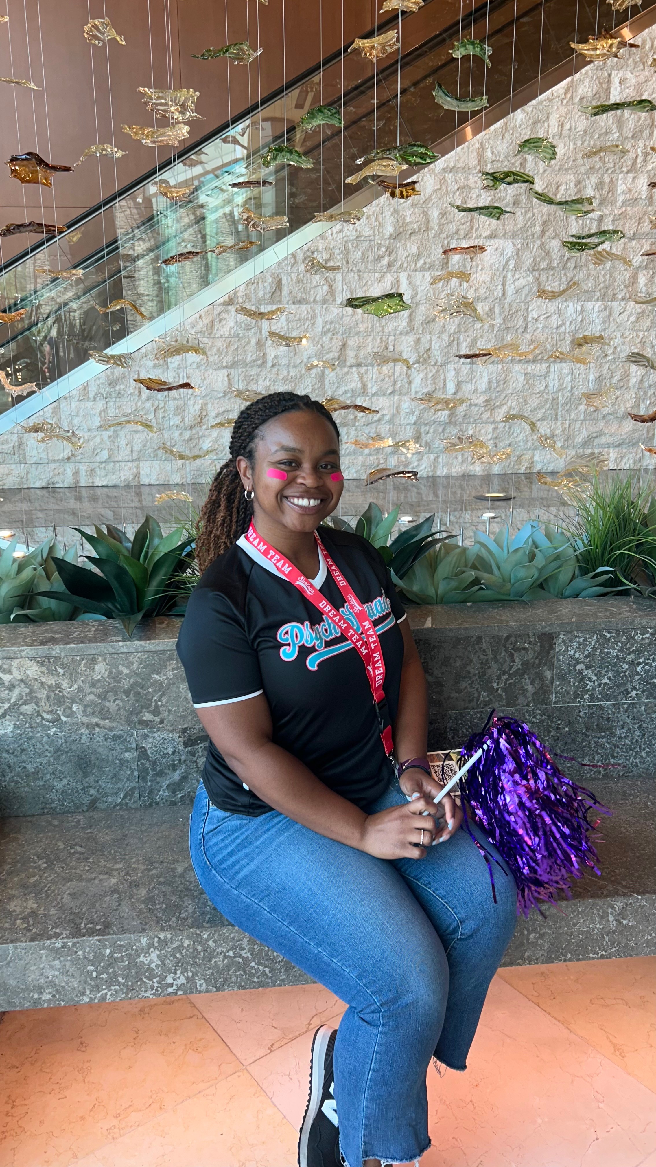Smiling woman sitting on steps holding purple pom-pom, wearing black shirt and jeans, outdoors with plants and stone wall behind.