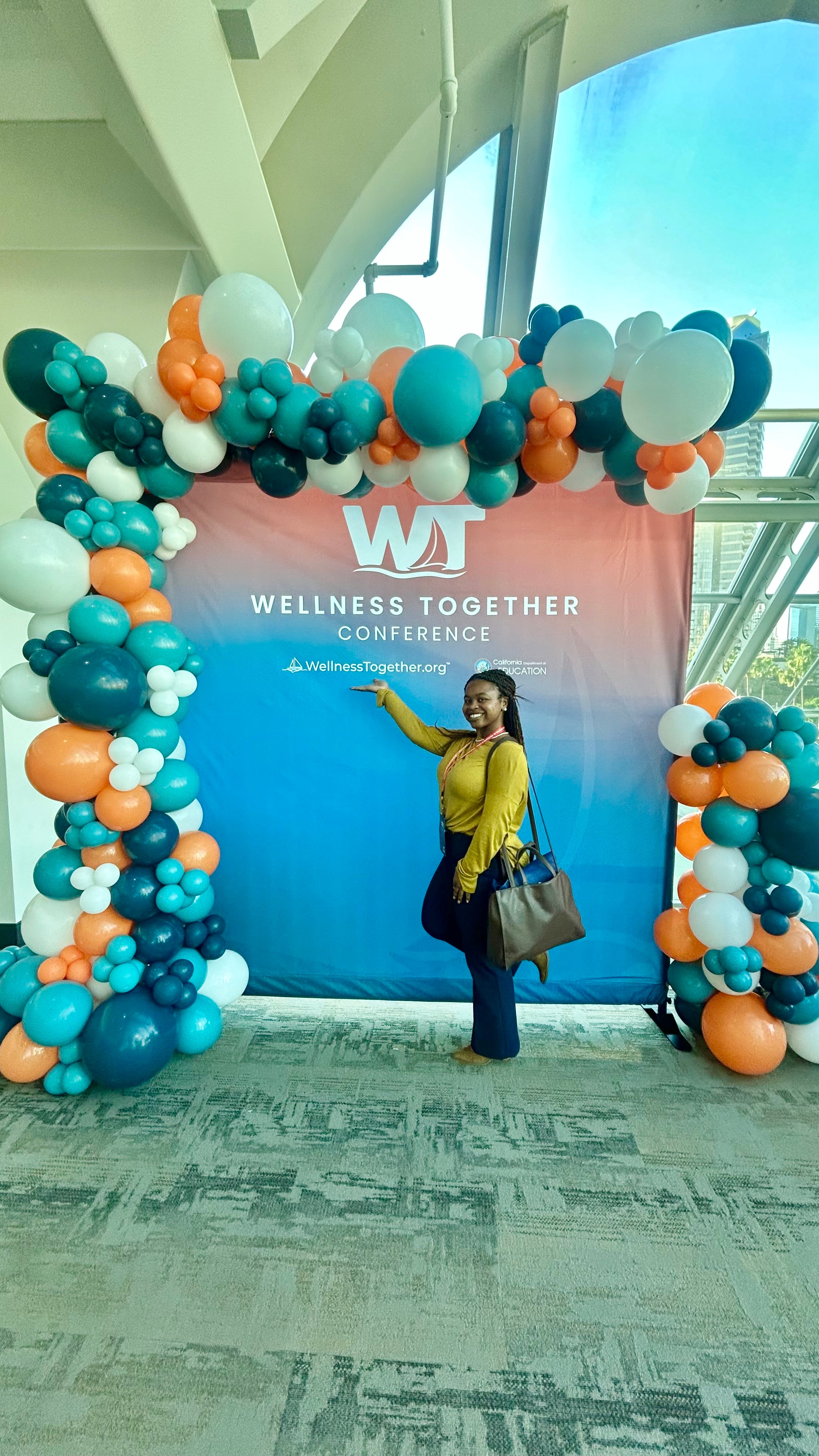 Person standing in front of a balloon arch at a wellness conference, with a large screen and window in the background.