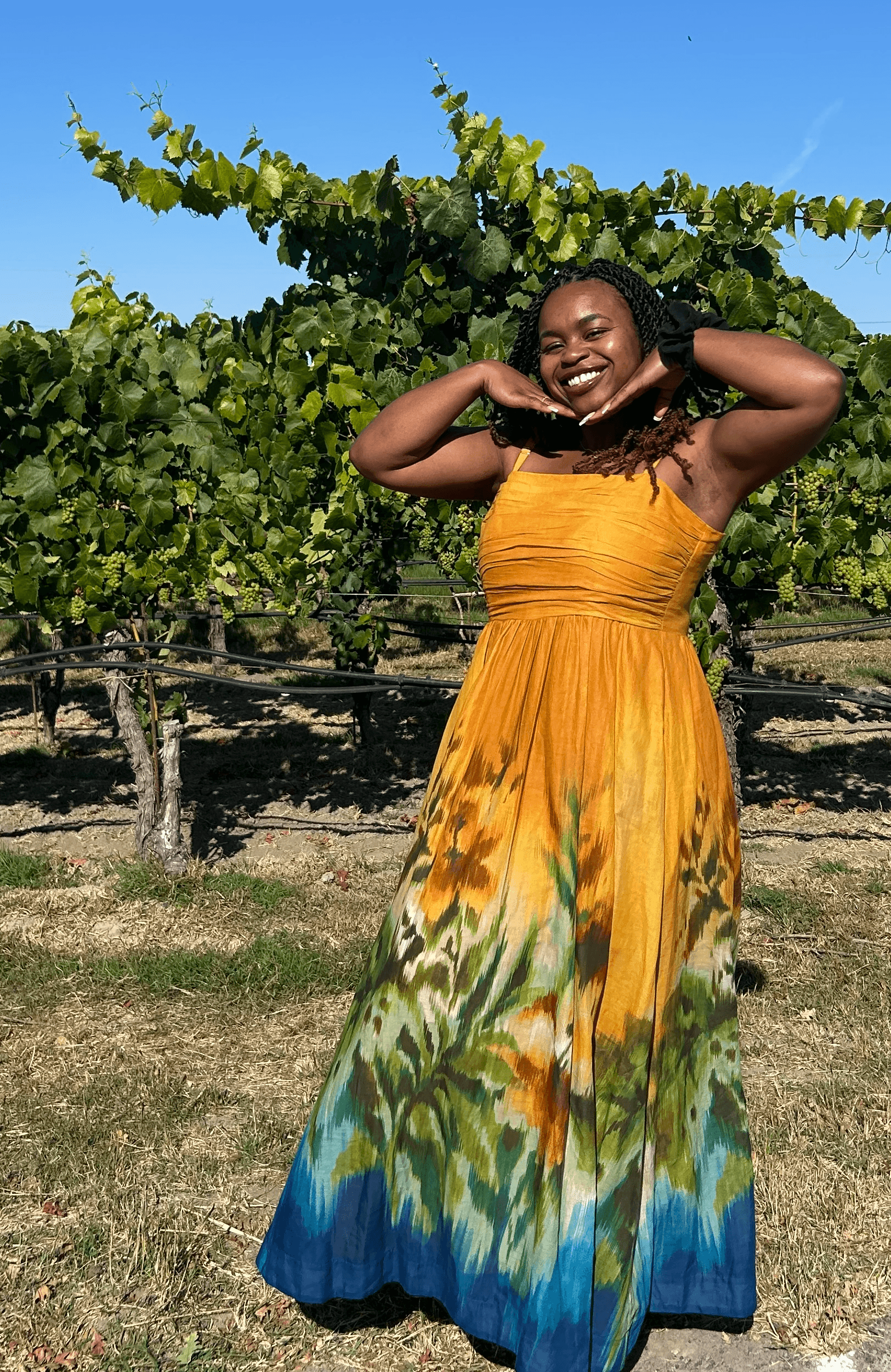 Smiling woman in a yellow and blue dress standing outdoors with green trees and clear blue sky behind her.