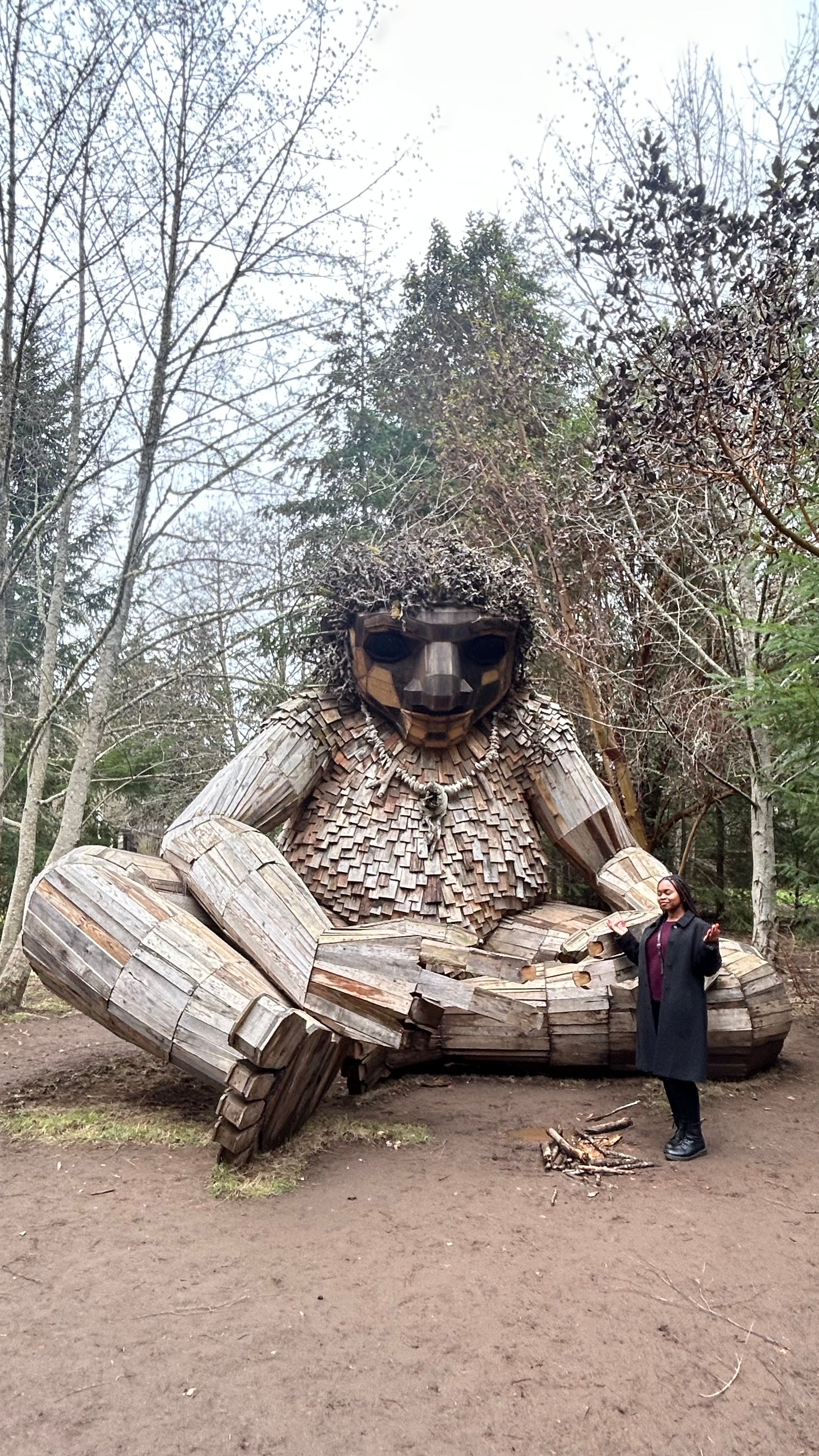 Large wooden sculpture of a bear with sunglasses, sitting outdoors among trees, with a person standing nearby for scale.