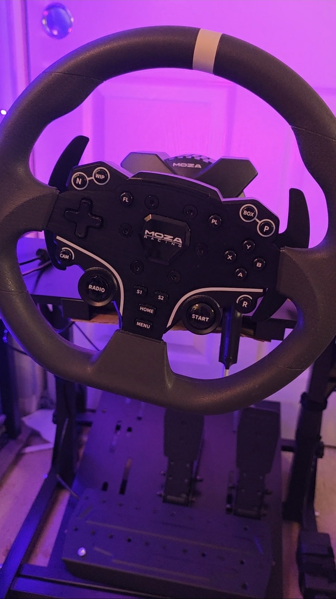 Steering wheel with gaming controls and pedals, illuminated by purple lighting.