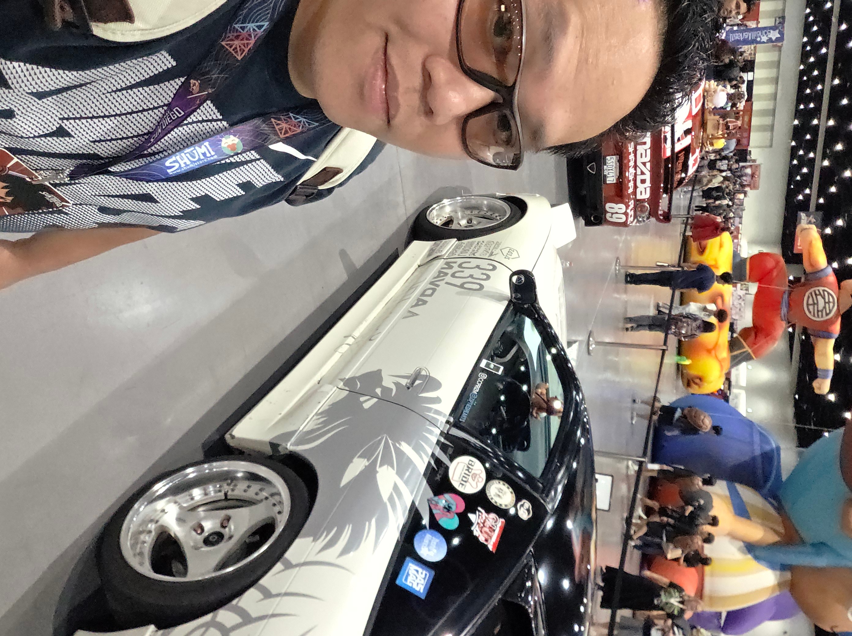 Person with glasses at a table near a white car model with black accents, in a busy indoor setting.