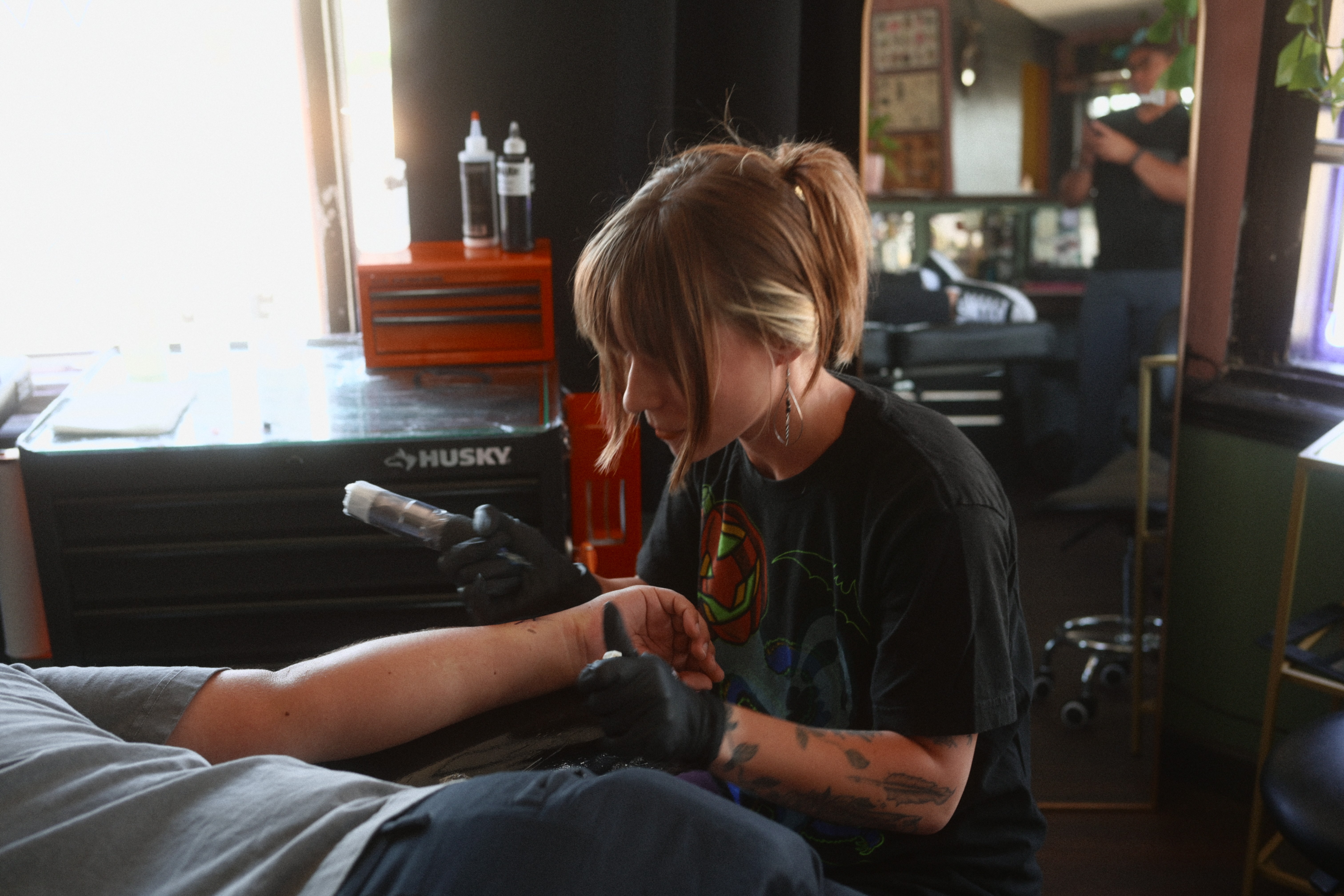 Person with long hair and tattoos sitting at a table, with a person’s arm visible, in a room with a window and various objects