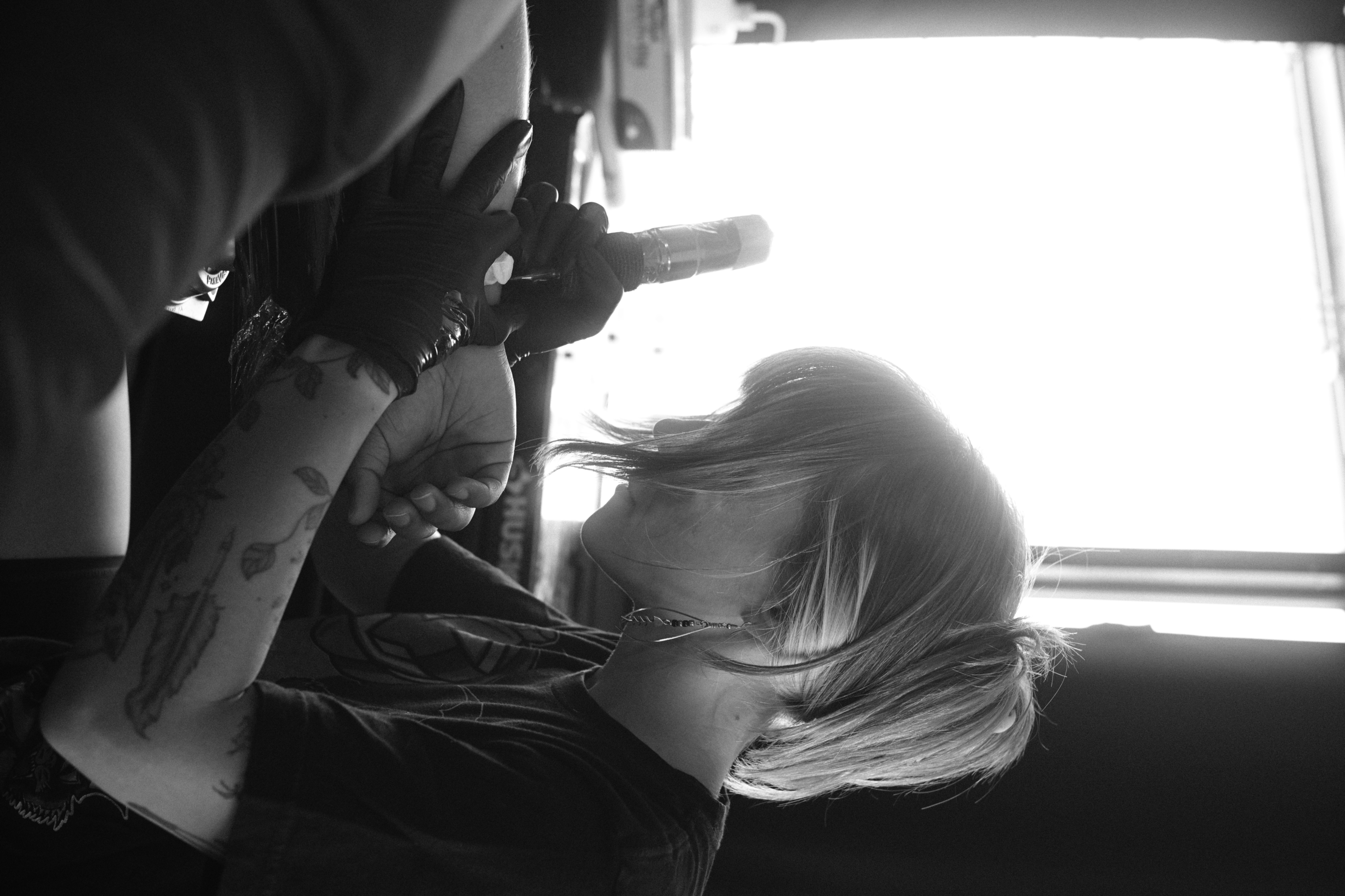 Person with tattoos and glasses holding a phone, with a window in the background, in black and white.