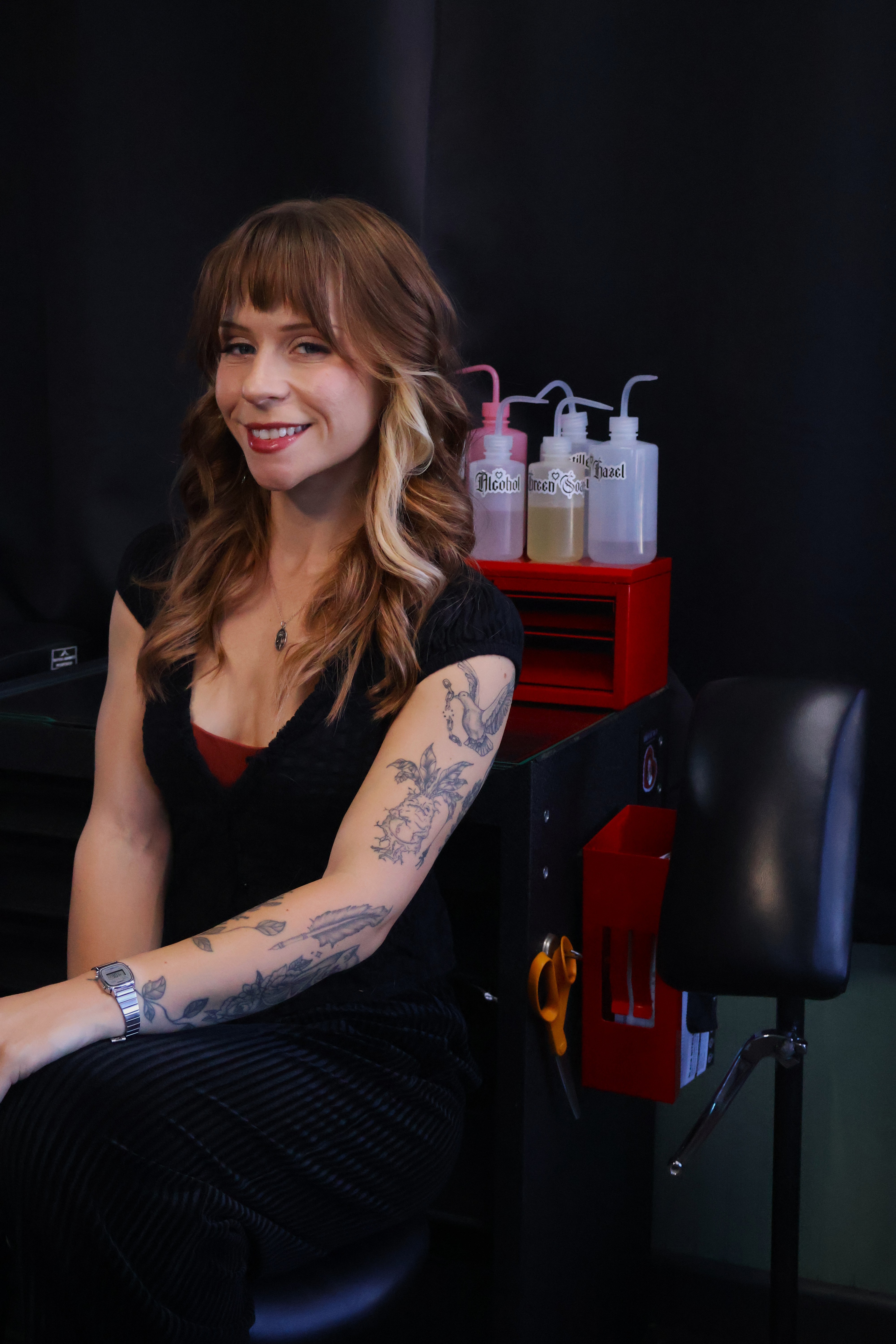 Woman with long wavy hair and tattoos on her arm, smiling, sitting in a chair with a dark background.