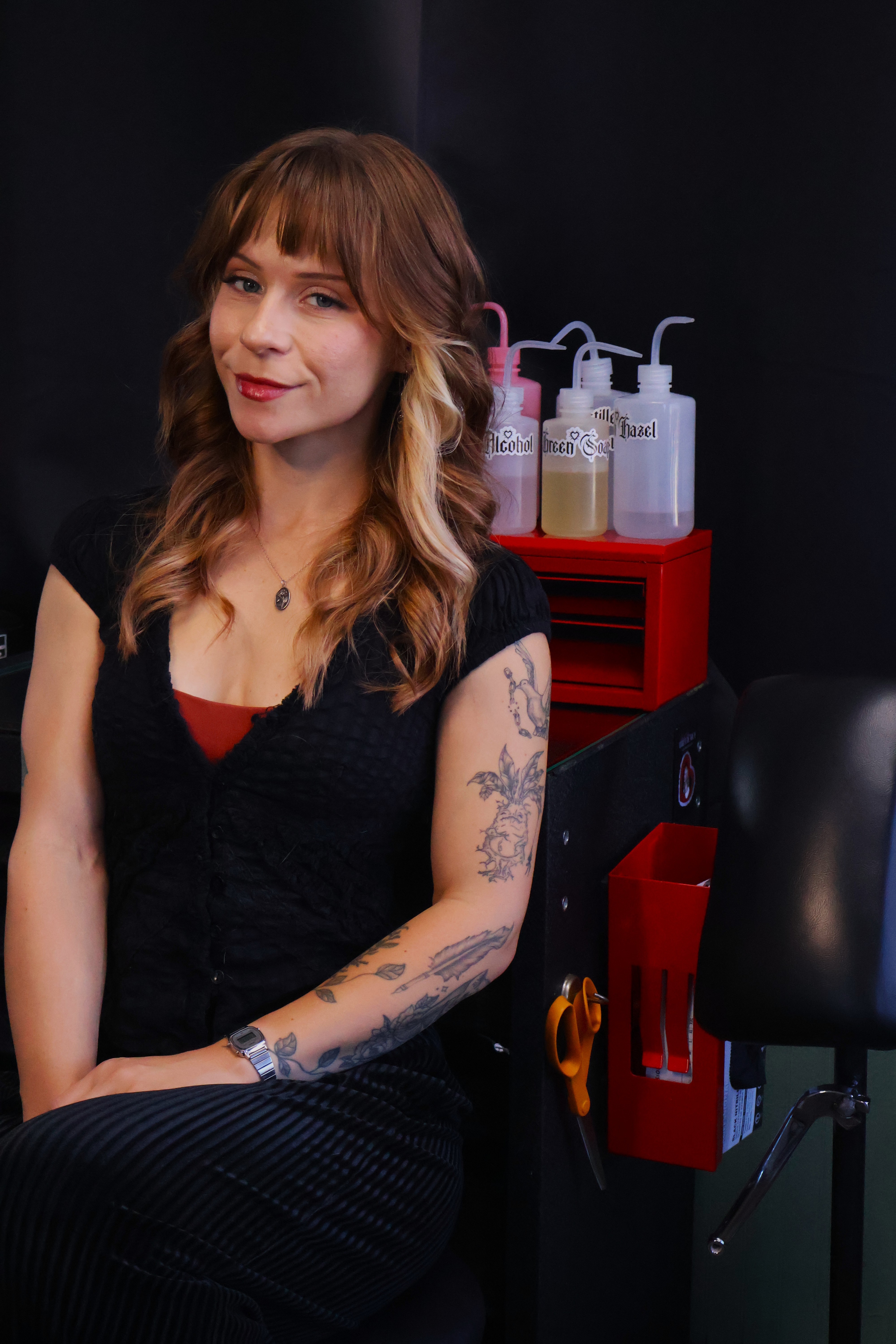 Woman with wavy brown hair and tattoos on her arm, wearing a black top, sitting in a salon or studio setting.