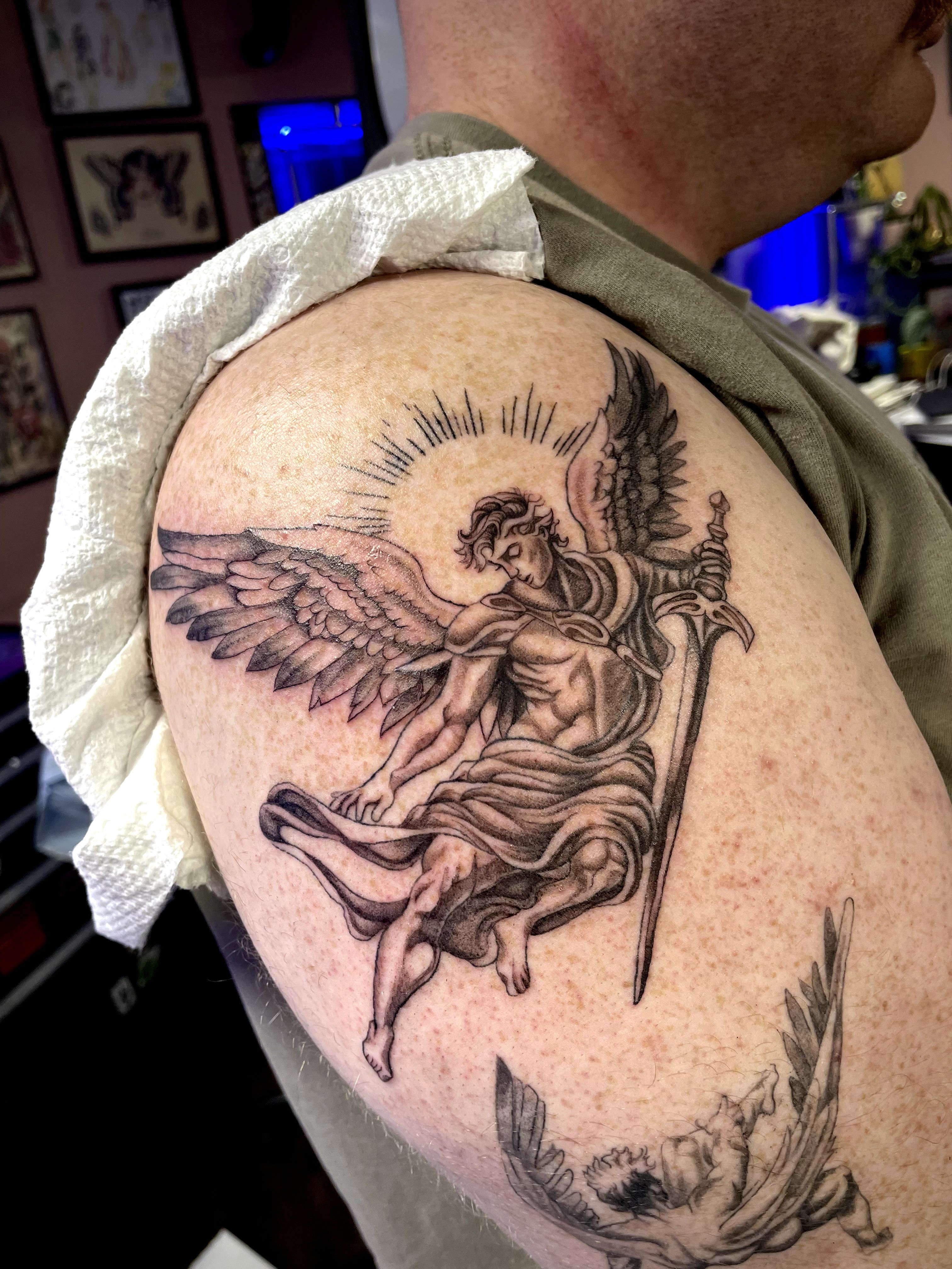 Tattoo of a winged humanoid figure with a halo, holding a spear, seated on a cloud, on person's upper arm.