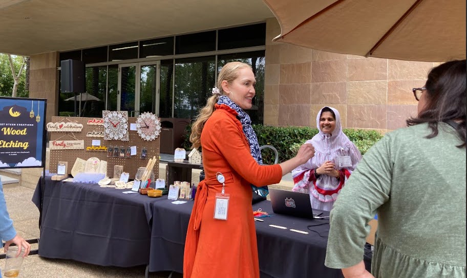 Woman in orange coat talking to a person at an outdoor market stall with jewelry and crafts, building in background.