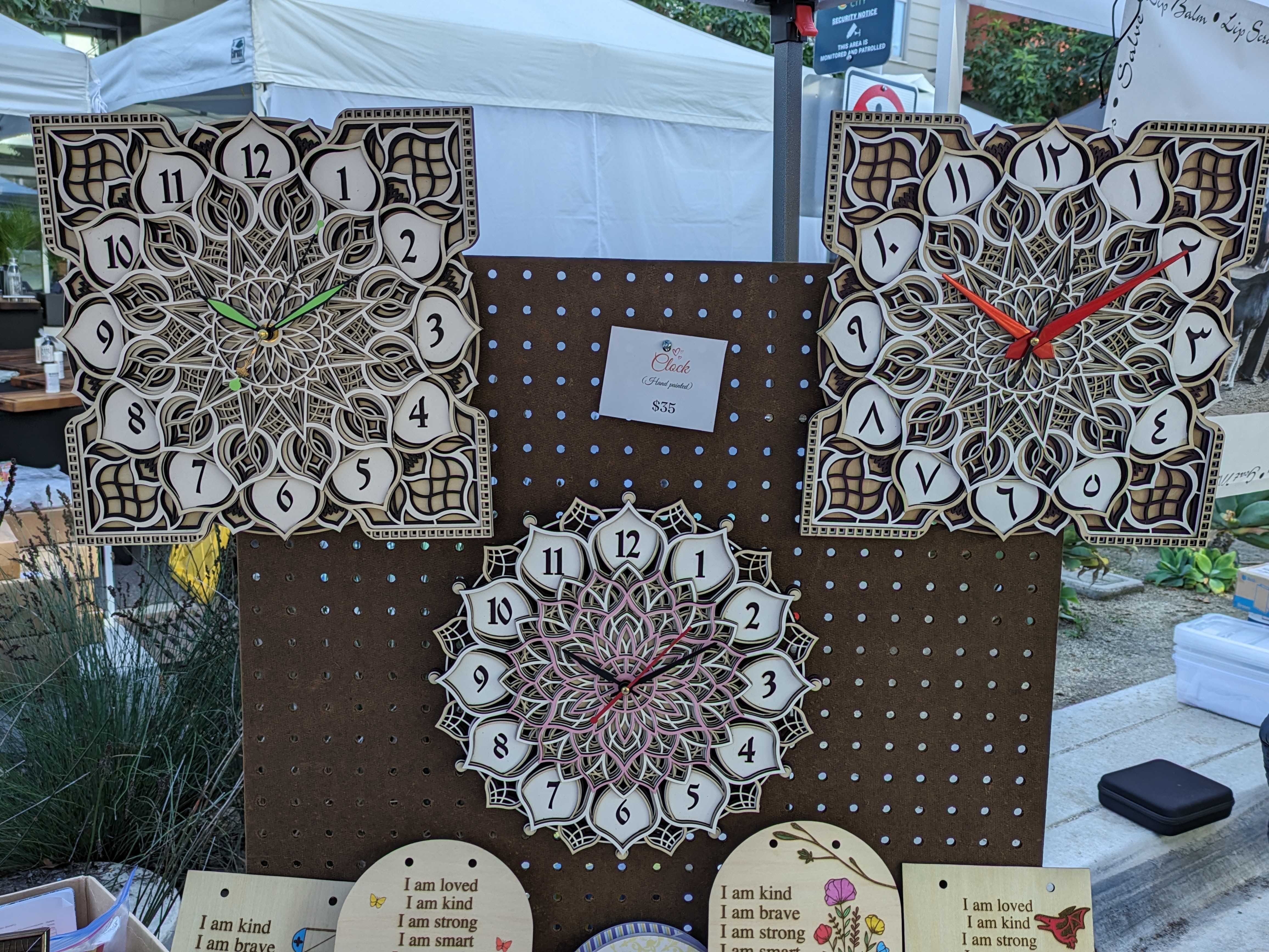 Three decorative clocks with intricate designs and different colored hands are displayed on a brown pegboard outdoors.