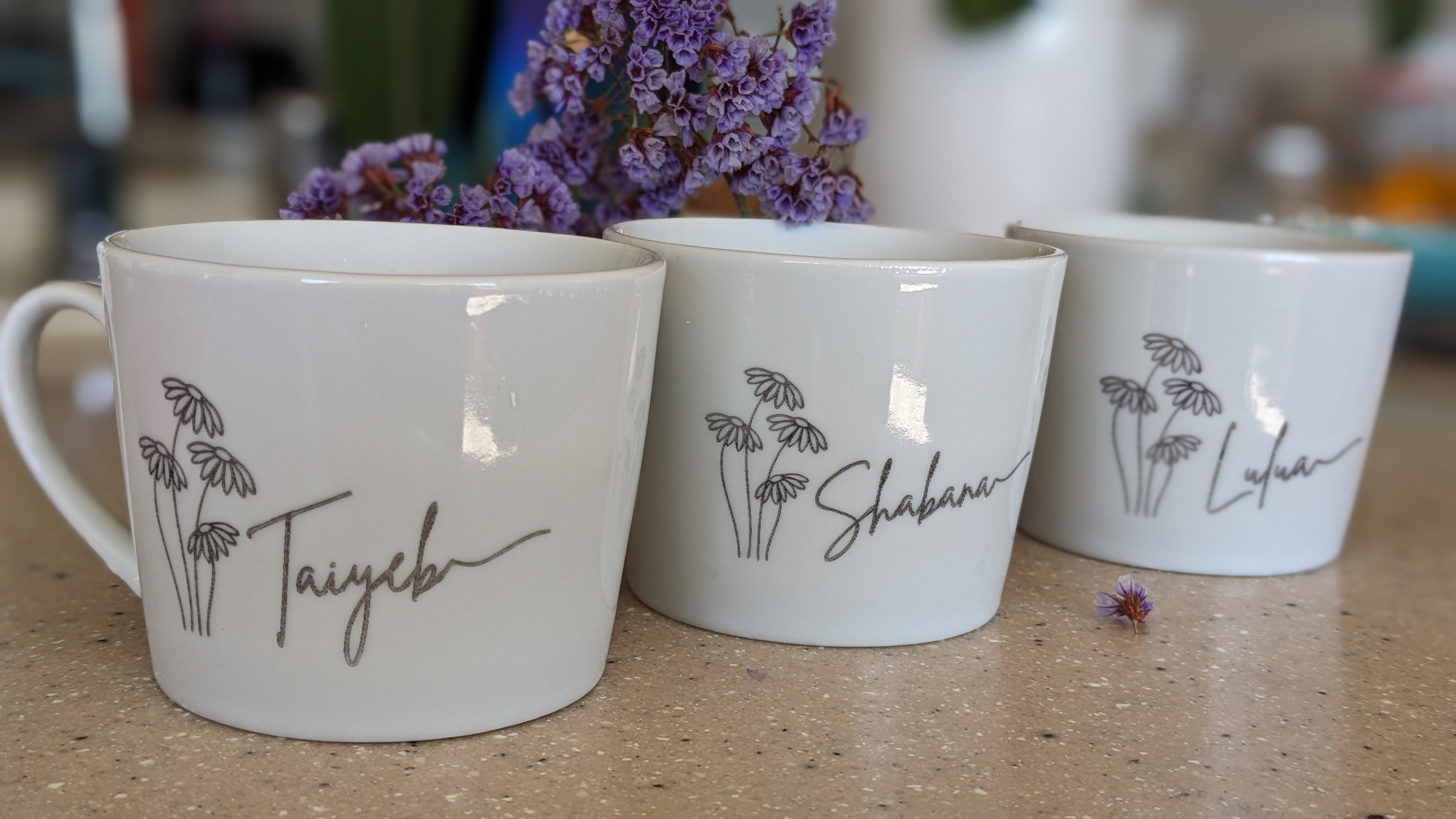 Three white mugs with plant illustrations and handwritten names, placed on a surface with purple flowers in the background.