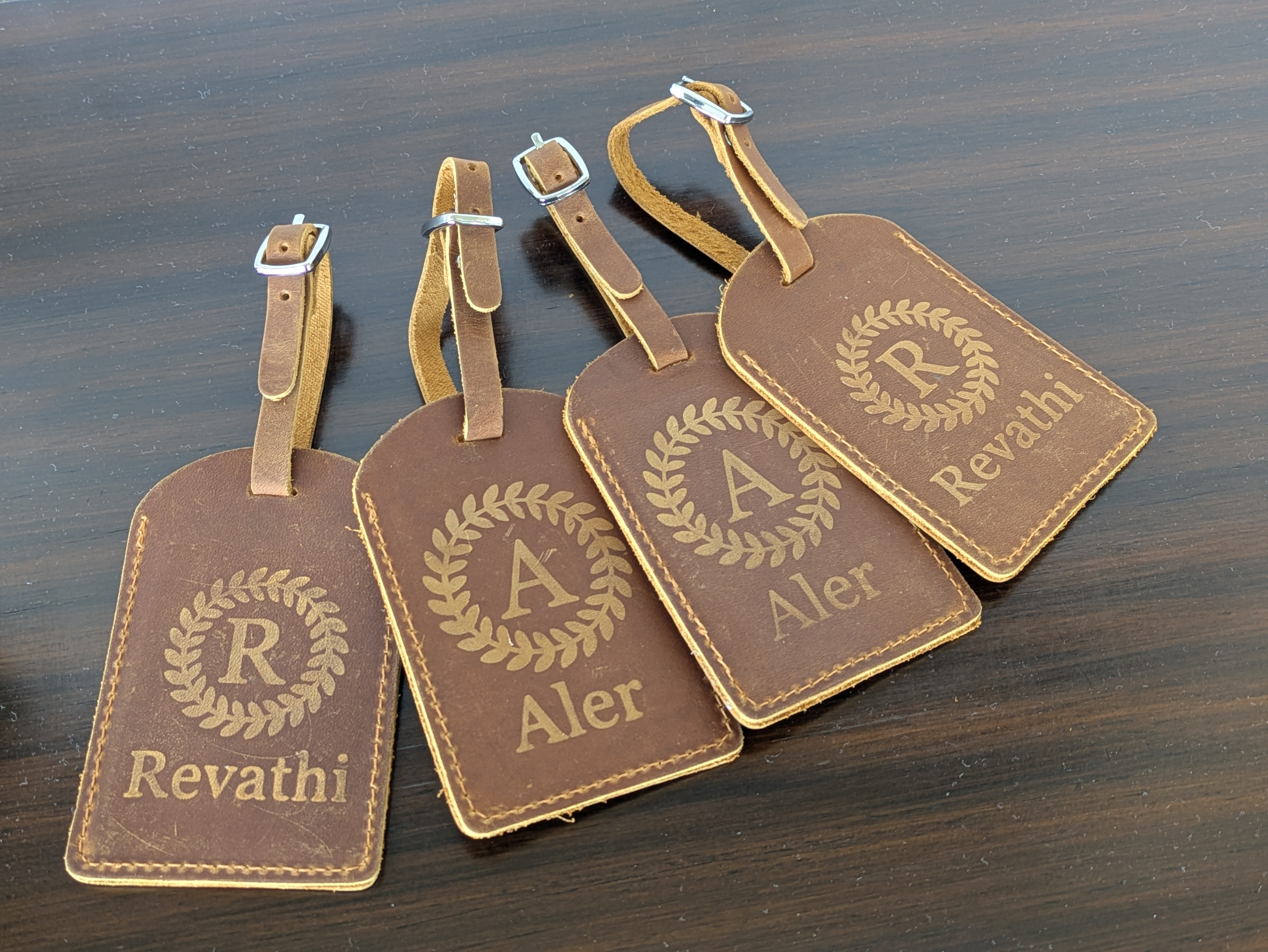 Four brown luggage tags with gold lettering and logos, labeled R, Aler, and Revatih, on a dark wooden surface.