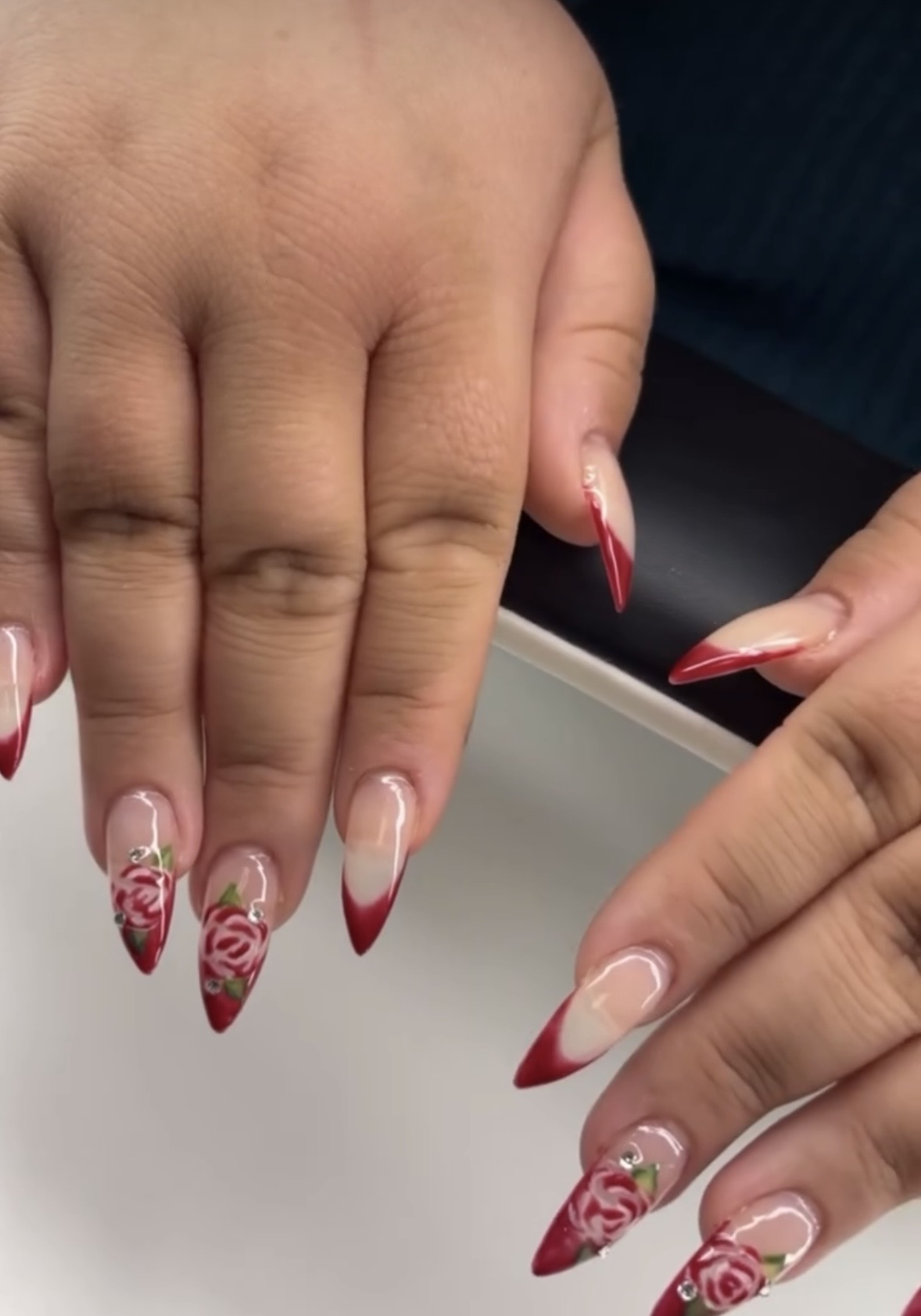 Two hands with decorated nails, featuring red and white designs, including floral and swirl patterns, on a dark background.