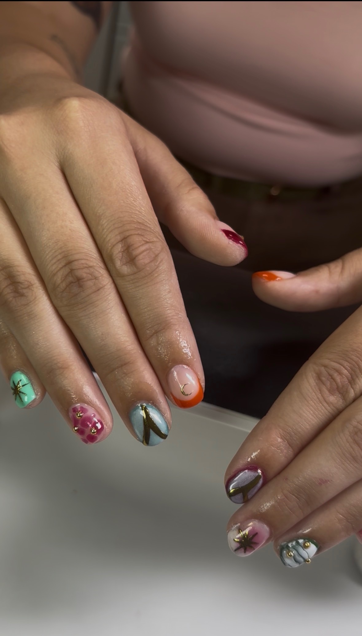 Close-up of hands with colorful, decorated fingernails, some with patterns and embellishments, resting on a surface.