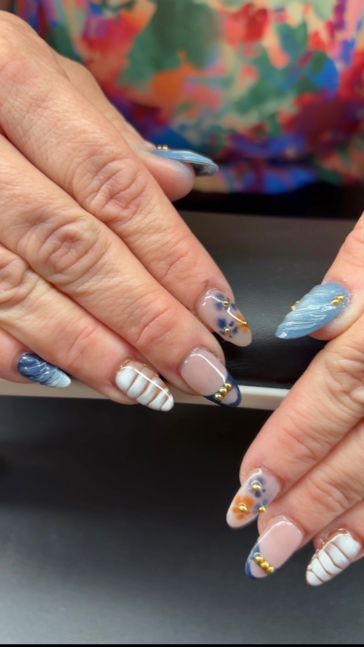 Close-up of hands with decorated nails, some with blue, white, and gold designs, on a dark surface.