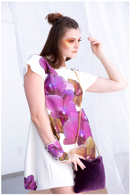 Young woman with brown hair in a top knot, wearing a white dress with purple floral patterns, holding a purple purse, standing against a white background.