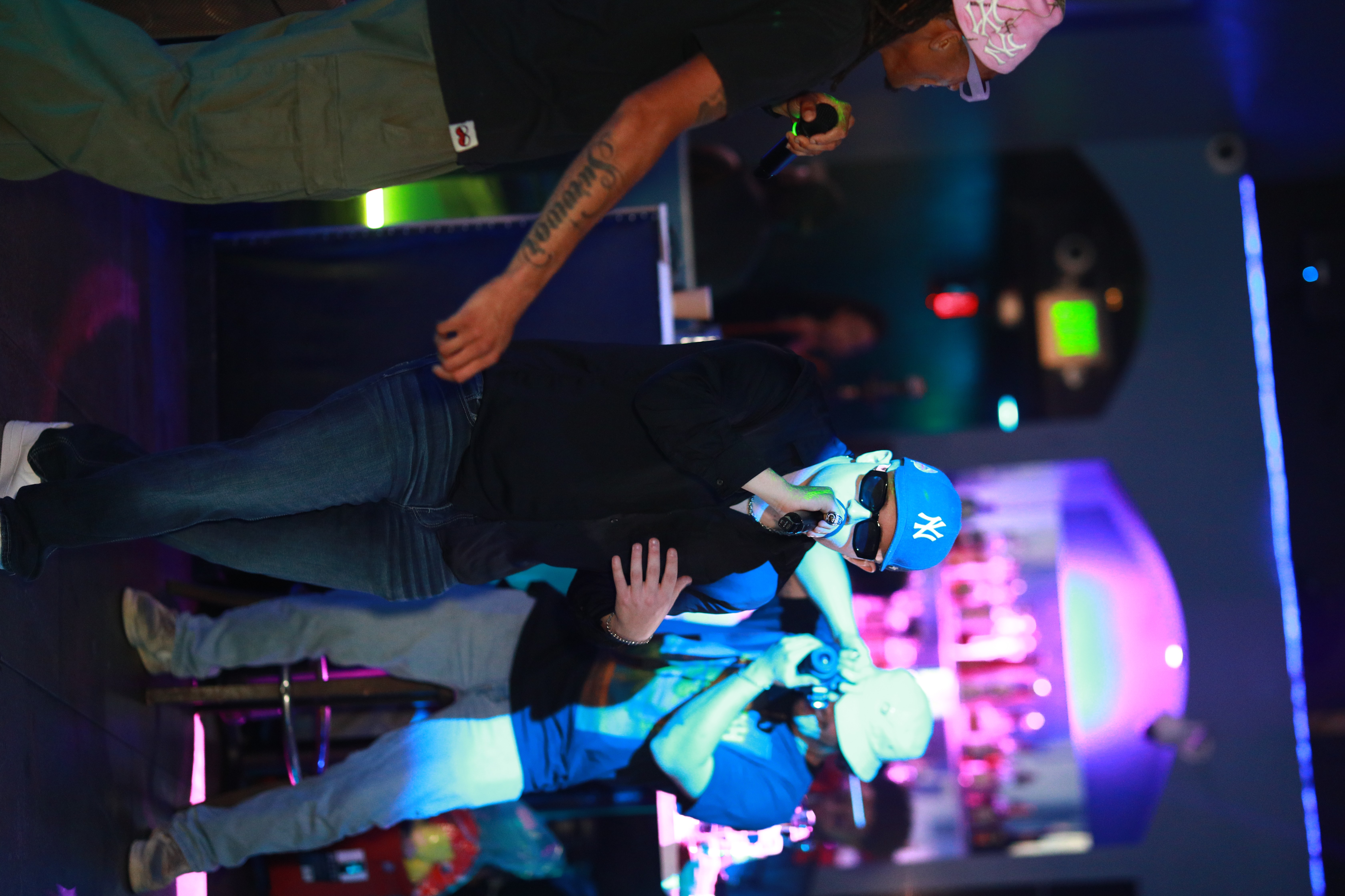 People in a nightclub with colorful lighting, some wearing hats, engaging in conversation or dancing, bar in background.
