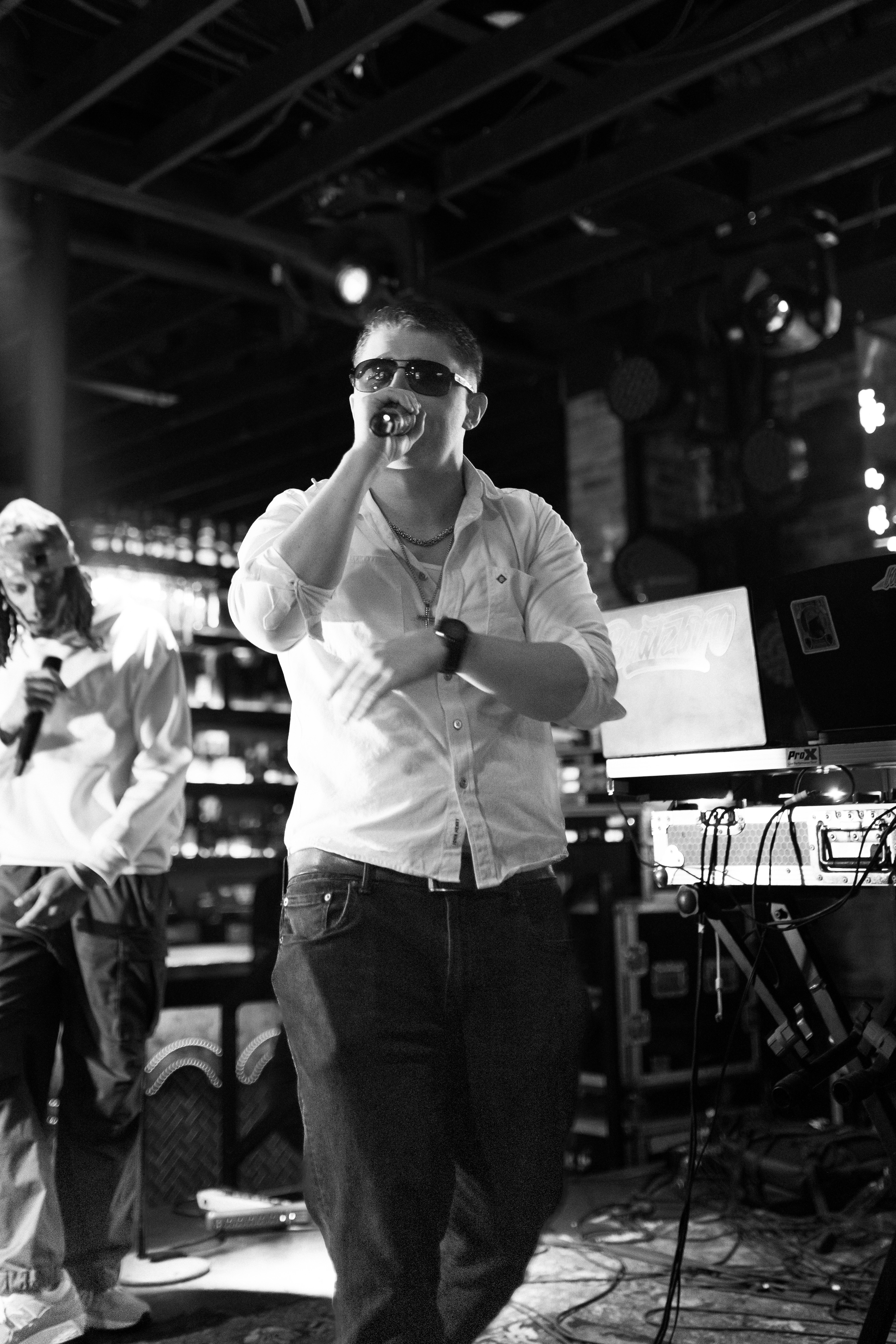 Man wearing sunglasses and a white shirt, holding a microphone, standing on stage with equipment behind him.