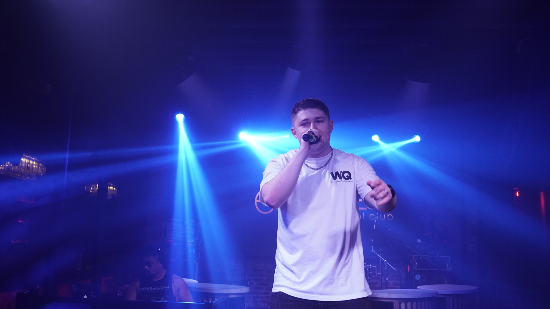 Person singing into microphone on stage with blue lighting and spotlights, holding a drink, wearing a white shirt.