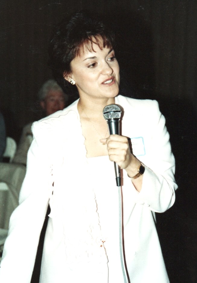 Woman with short dark hair in a white blazer holding a microphone, speaking at an event.