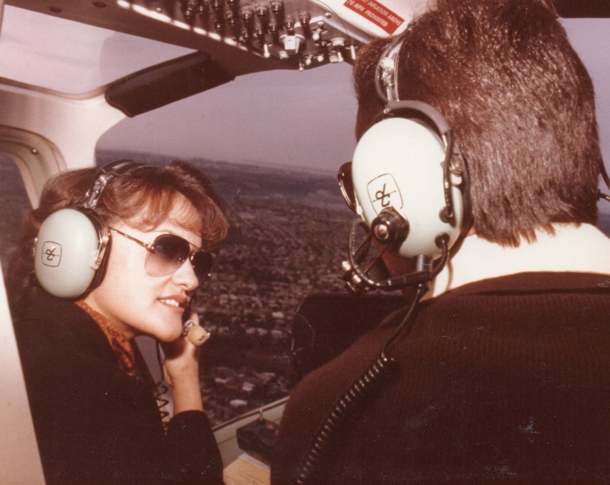 Two people wearing headsets inside a helicopter, with a cityscape visible through the window.