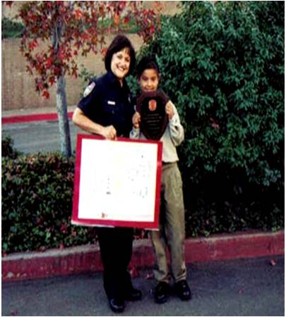 Two children standing outdoors, one holding a certificate and the other holding a plaque, with trees and bushes in background.