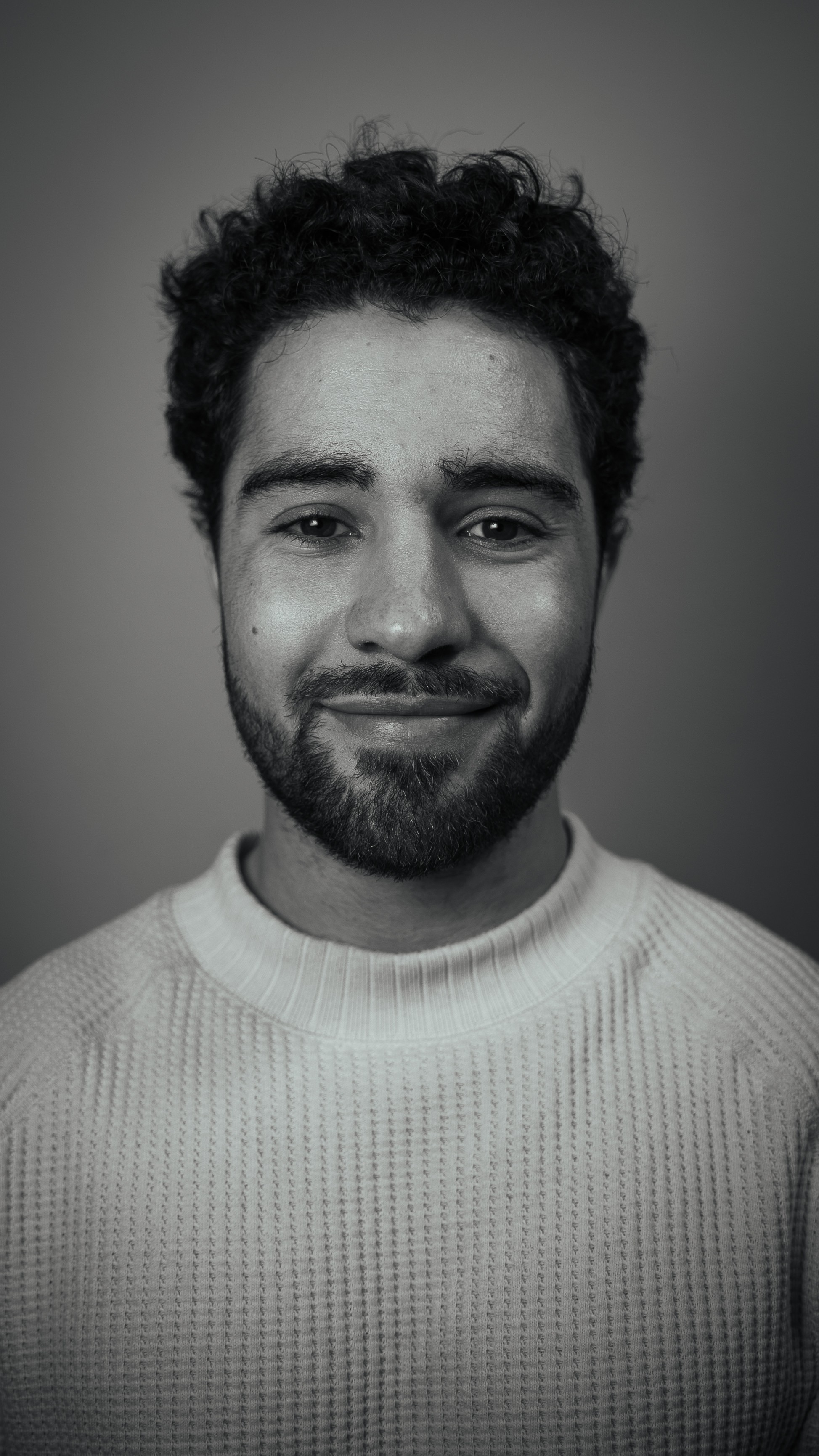 Young man with curly hair and beard smiling, wearing a light-colored sweater, against a plain background.