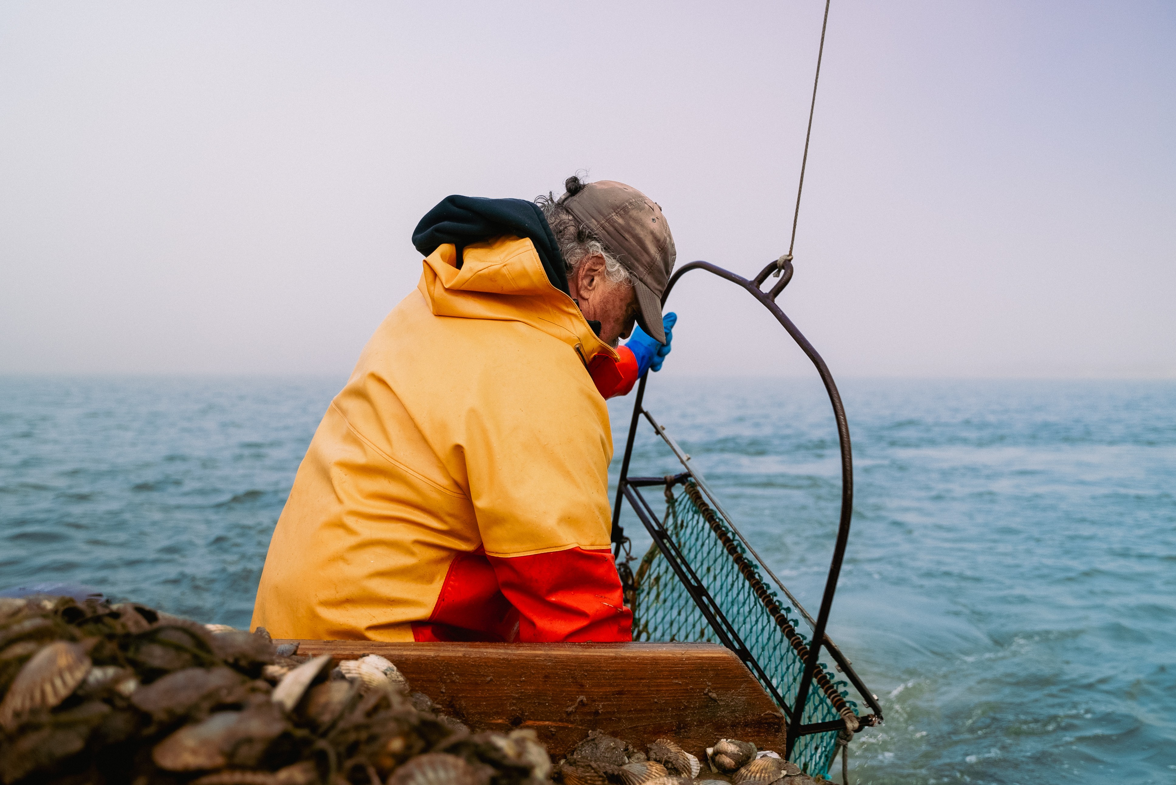 Person in yellow and red jacket sitting on rocks near water, looking down at the sea, with a metal structure nearby.