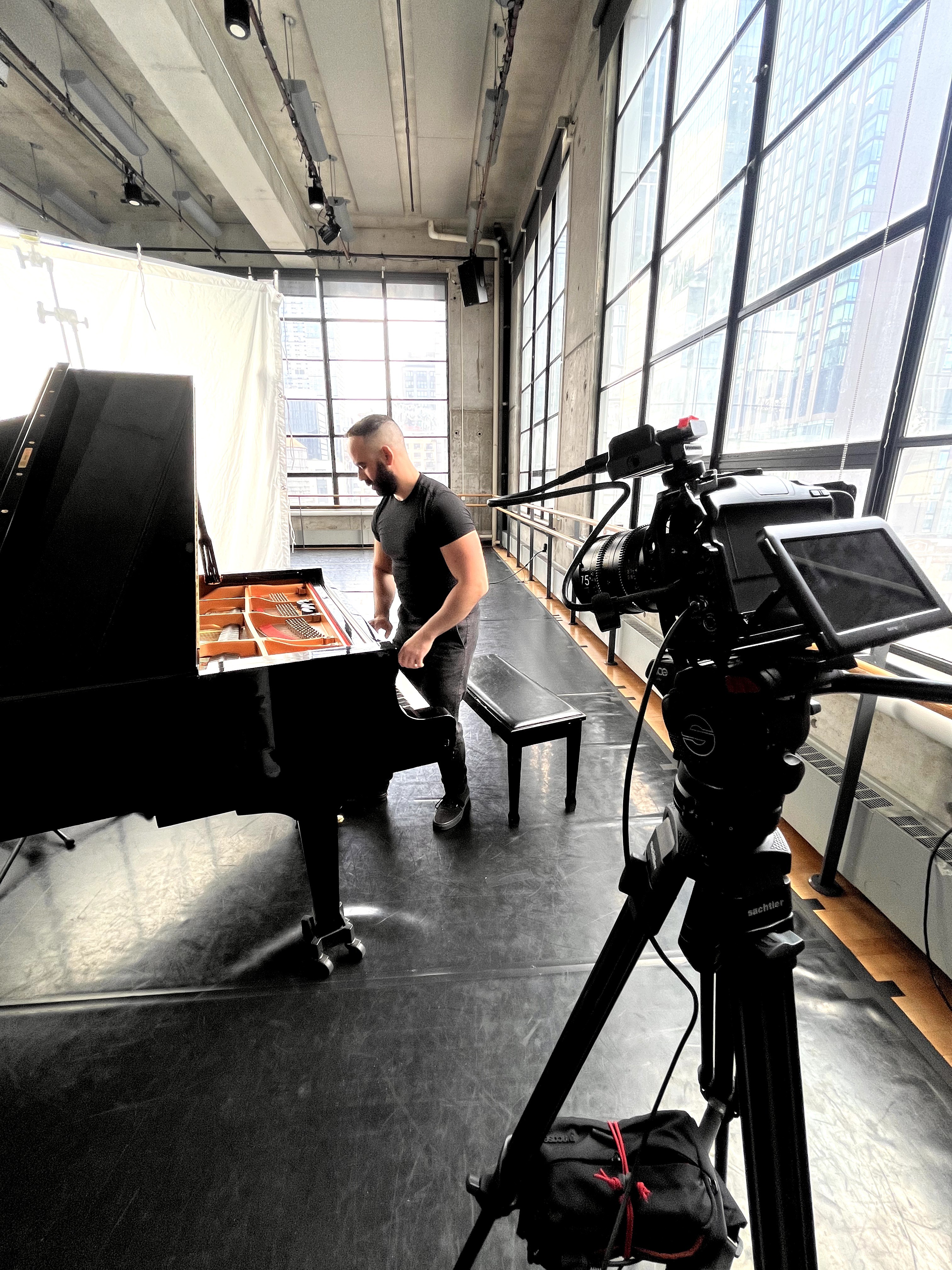Person playing grand piano in a bright studio with large windows, camera on tripod recording the scene.