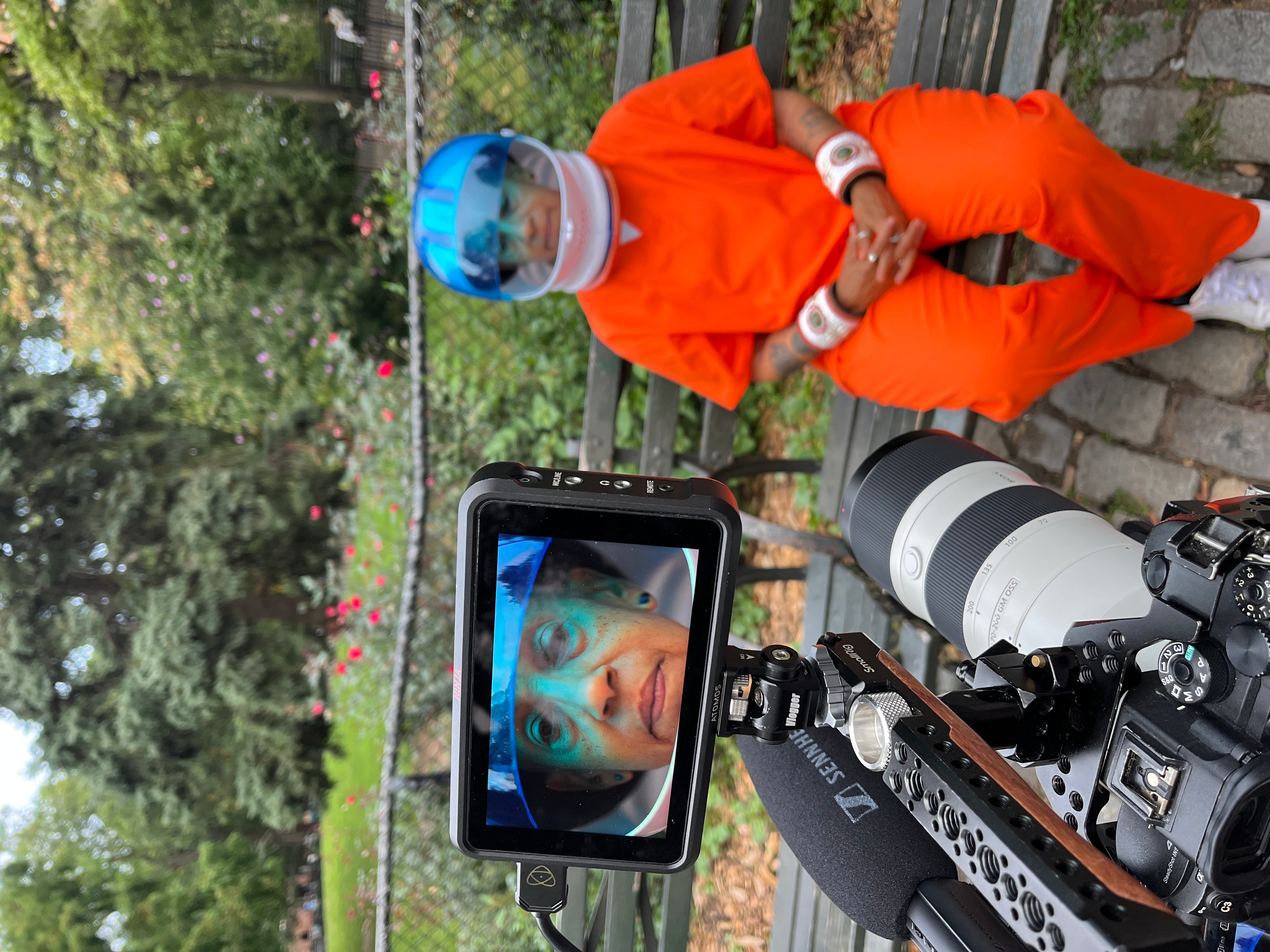 Child in orange outfit and blue helmet sitting on a bench, looking at a camera with a screen showing their face, outdoors.