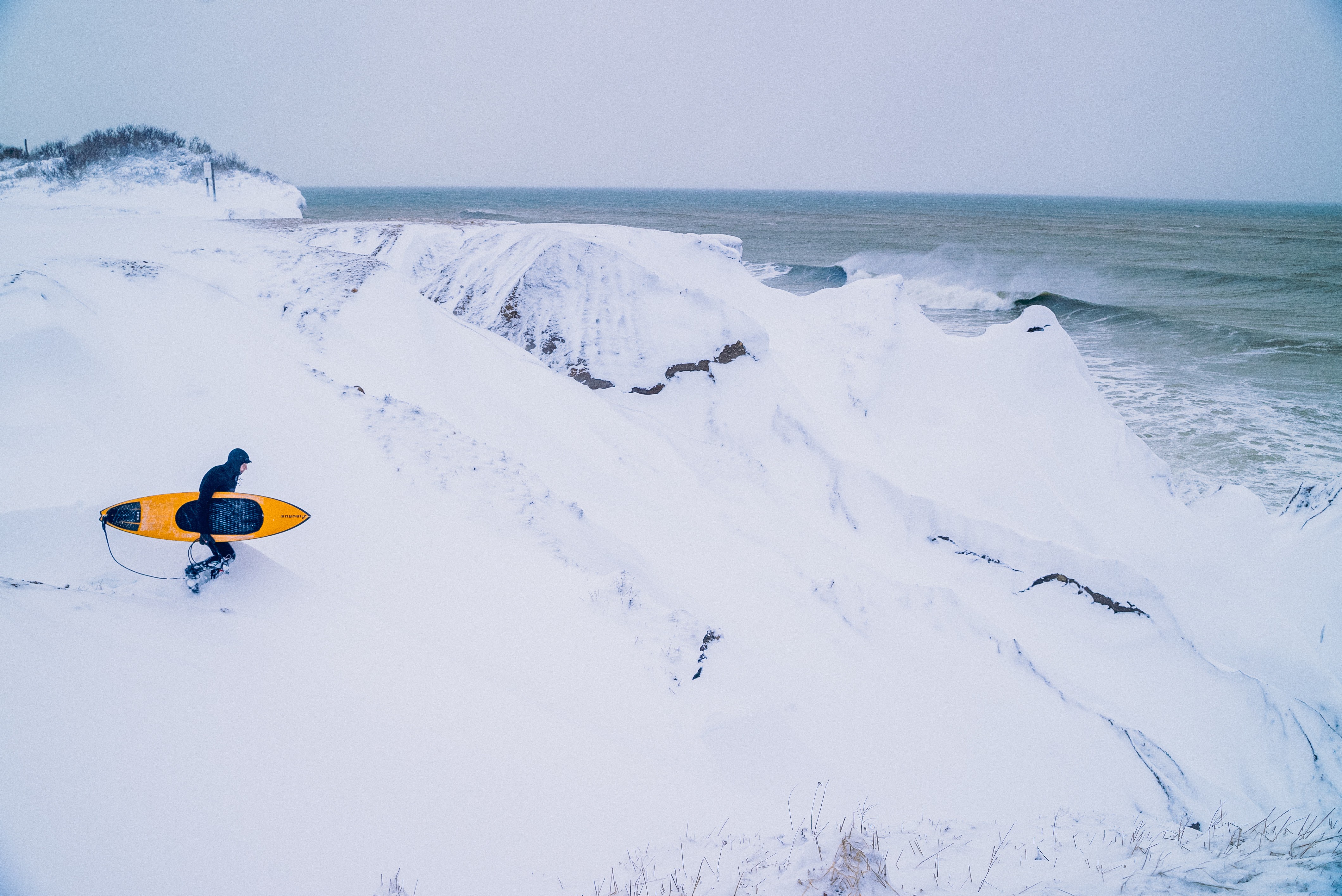 Person with a surfboard walking on snowy beach near ocean waves, overcast sky, snow-covered landscape, 20 words