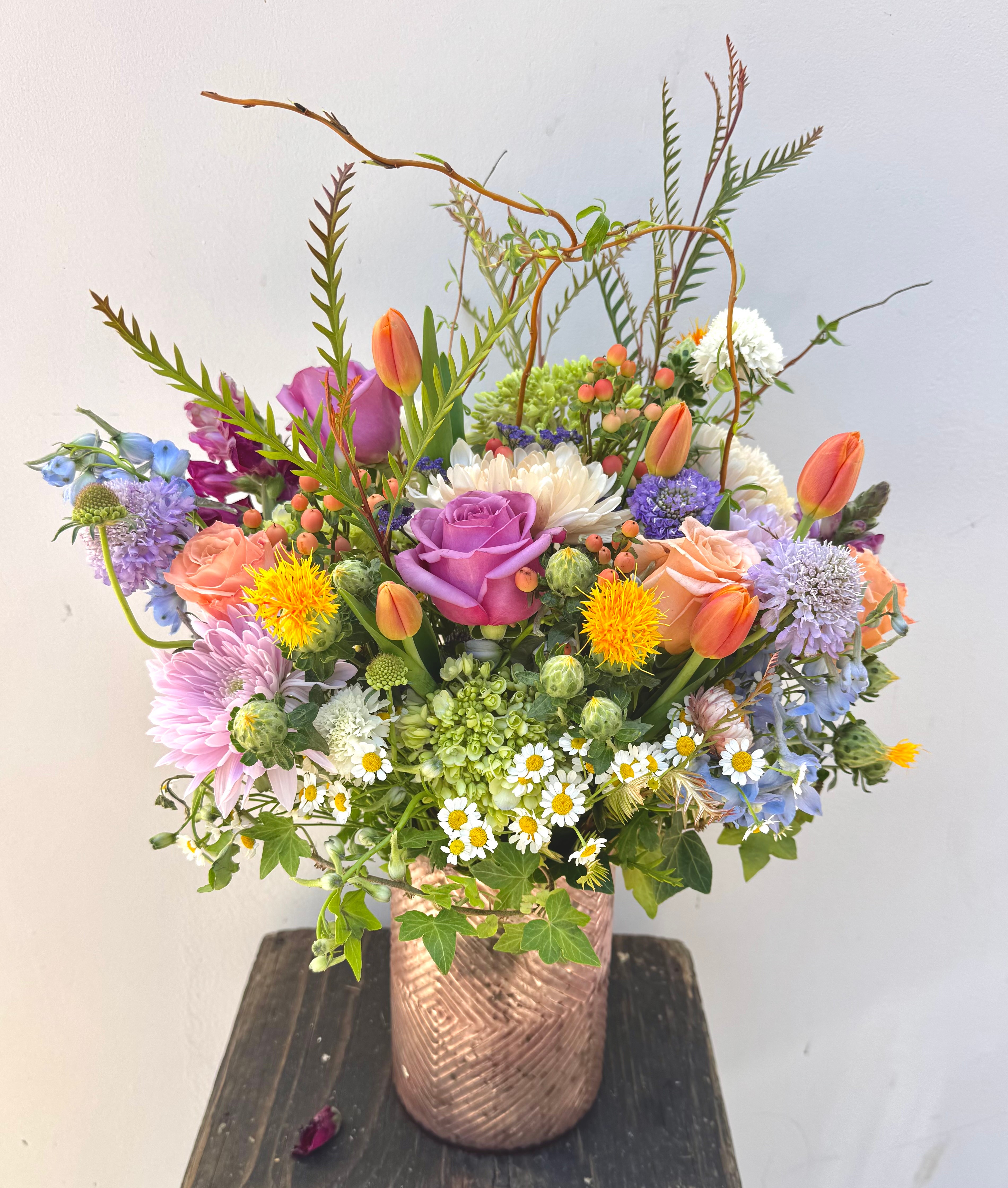 Colorful flower arrangement in a copper-colored vase with various flowers including tulips, daisies, and roses, on a dark surface.