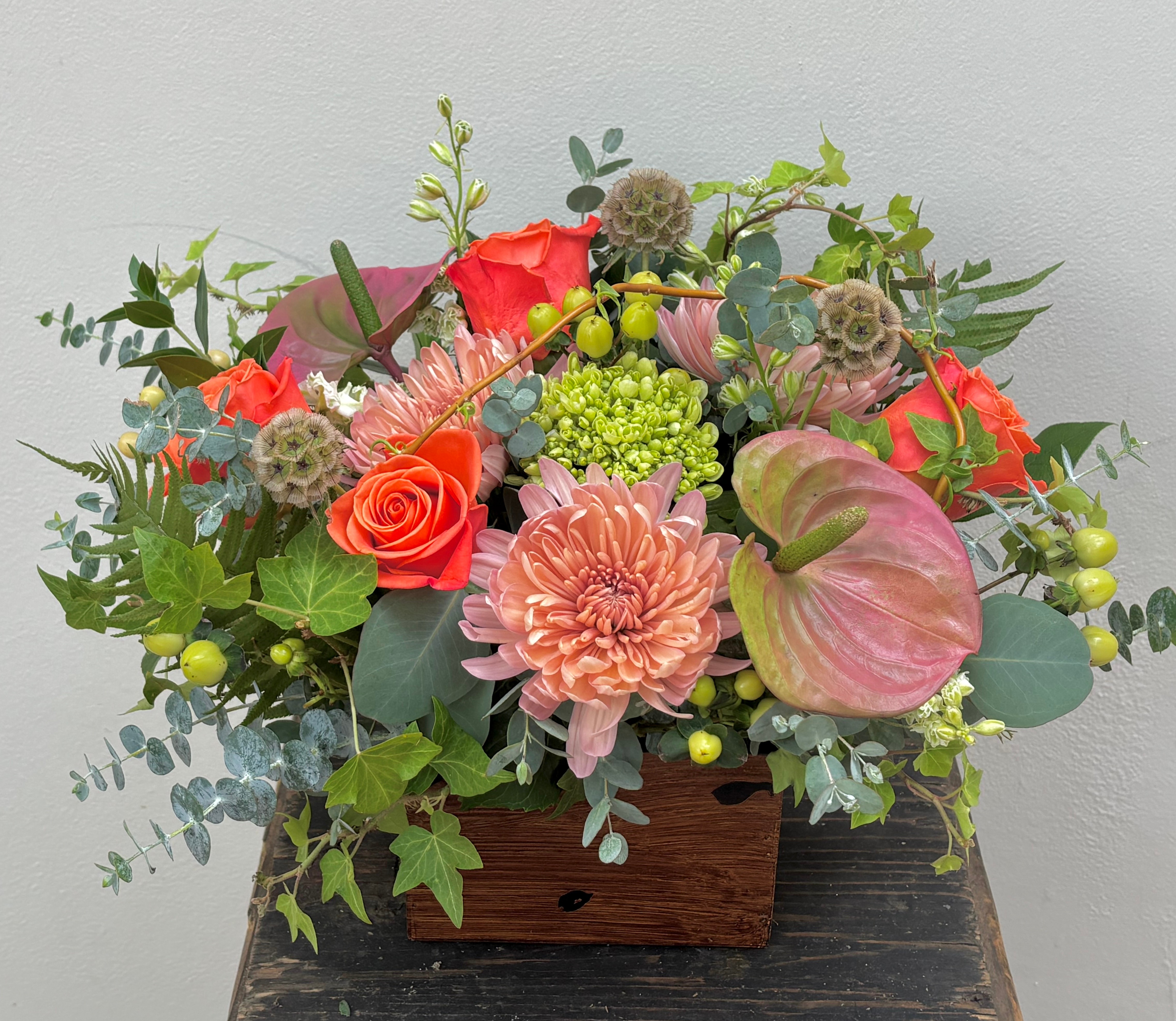 Arrangement of colorful flowers and greenery in a wooden box on a dark surface against a light background.