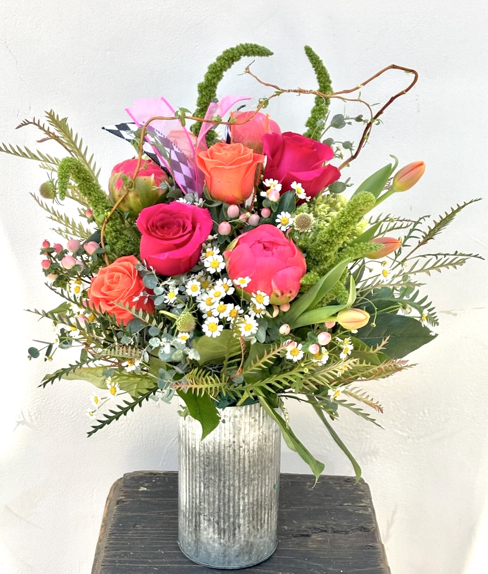 A colorful flower arrangement with roses, daisies, and greenery in a silver vase on a wooden surface.
