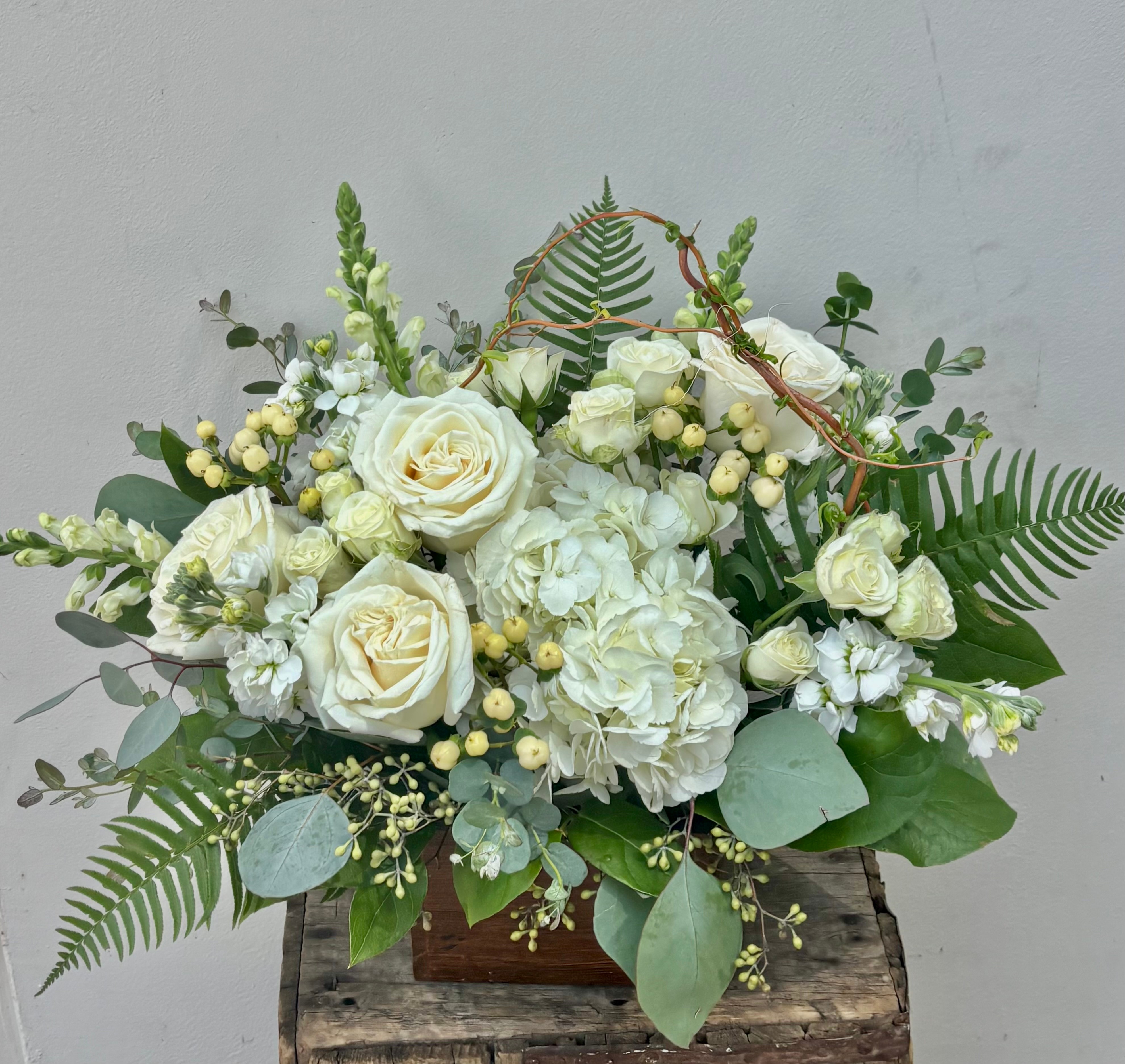 Arrangement of white roses, hydrangeas, and greenery in a wooden box on a rustic surface.