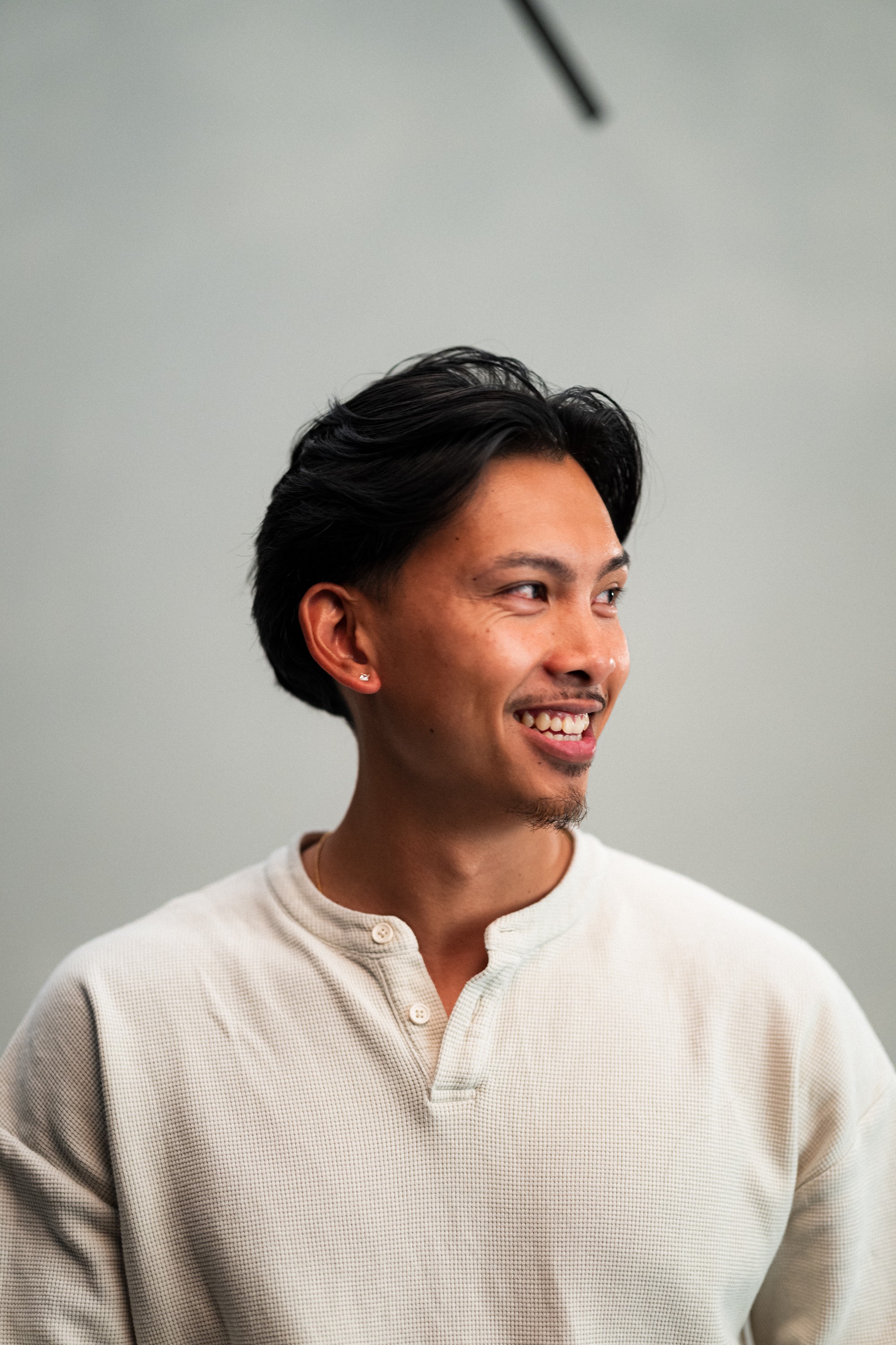 Smiling man with dark hair wearing a light-colored shirt, looking to the side, against a plain background.