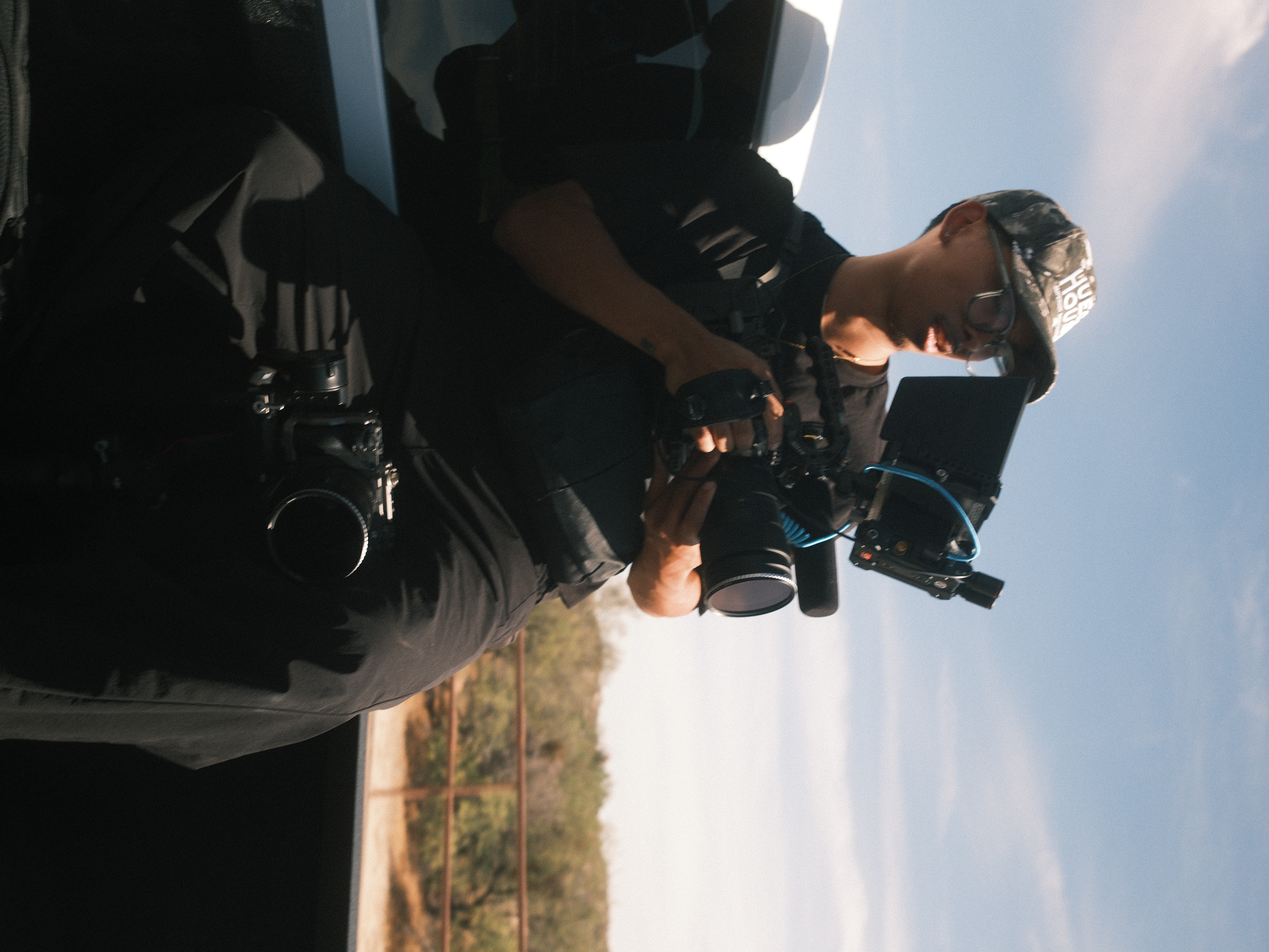 Person holding a camera with a camera rig, outdoors with a blue sky and clouds, side view.