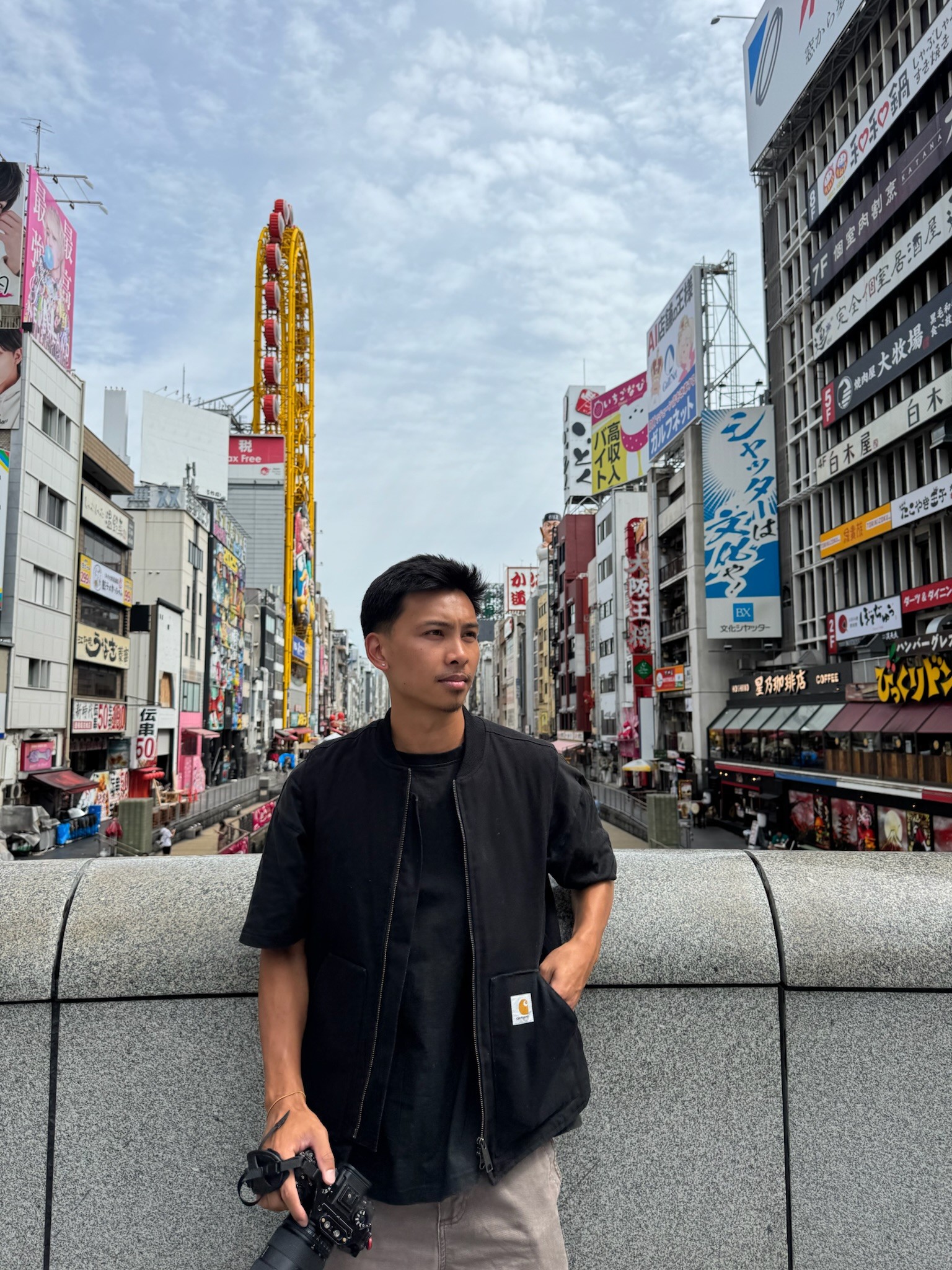 Young man standing in city street with tall buildings and colorful signs, holding a camera, in daytime.