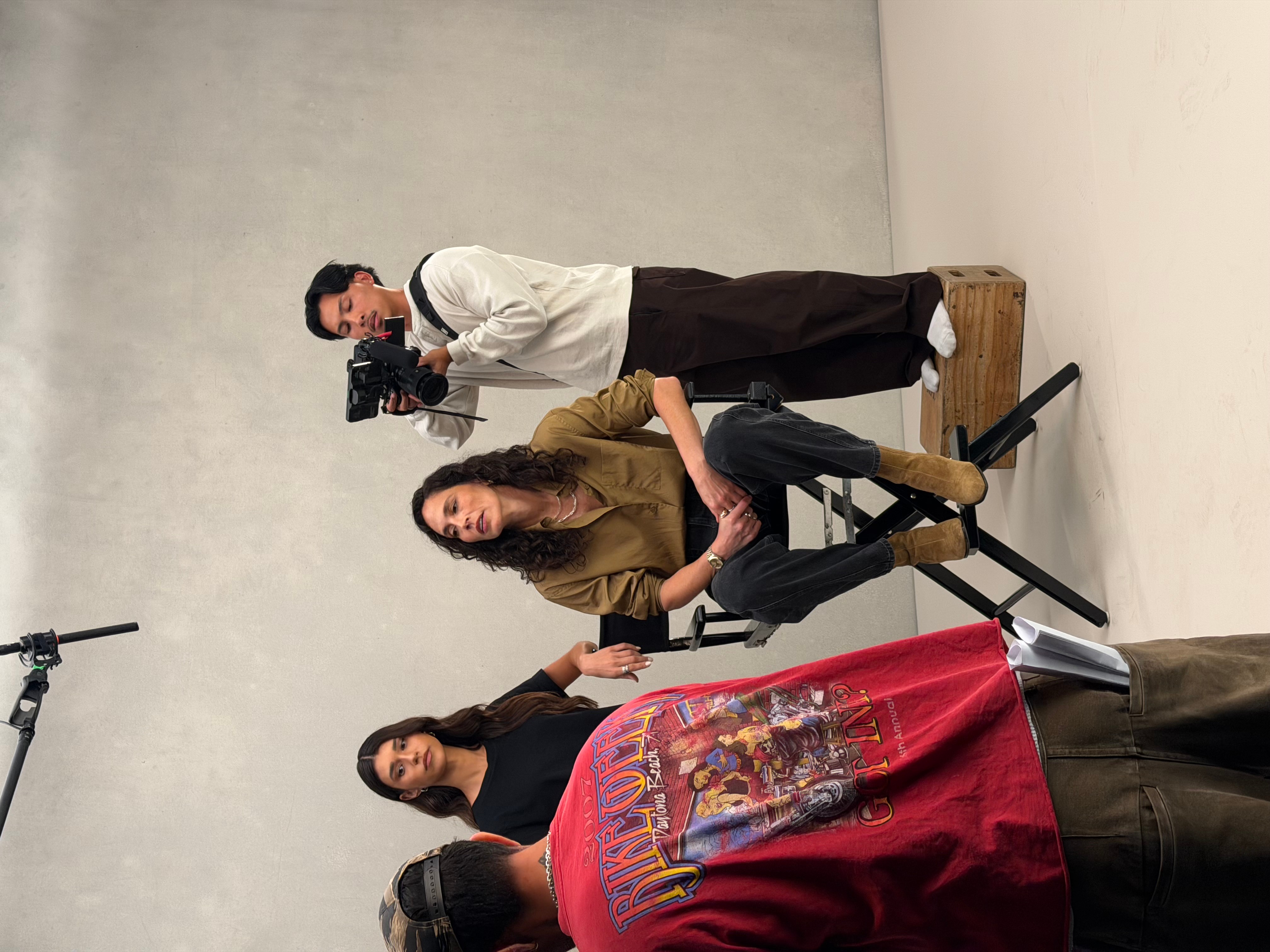 Three people in a studio, one person holding a camera, two seated, one standing, with a red cloth and a person in a red jacket in the foreground.