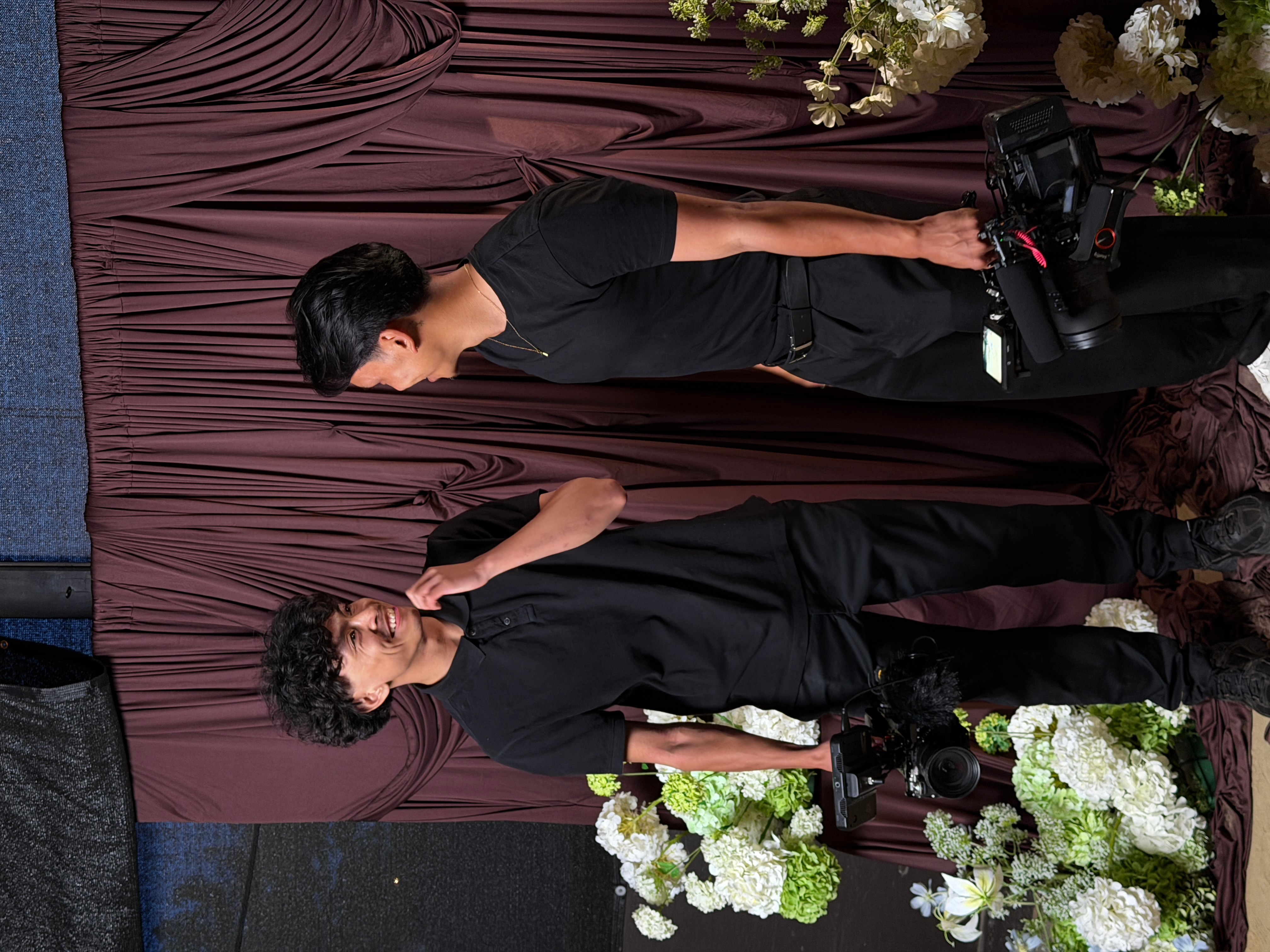 Two women standing in front of a brown curtain with flowers, one holding a camera, engaged in conversation.