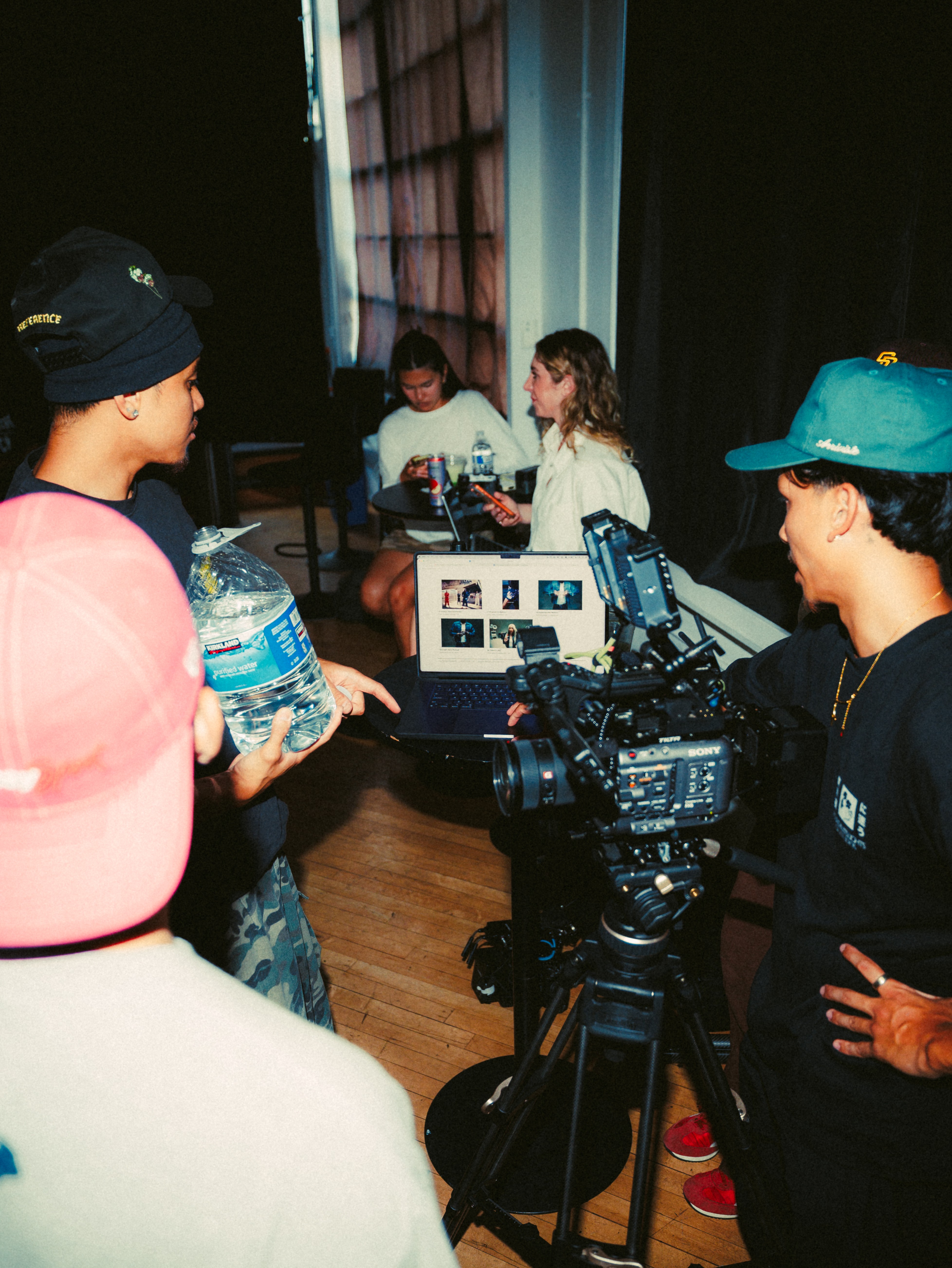 People in a room with a camera, two women sitting at a table, and others standing around, engaged in conversation.