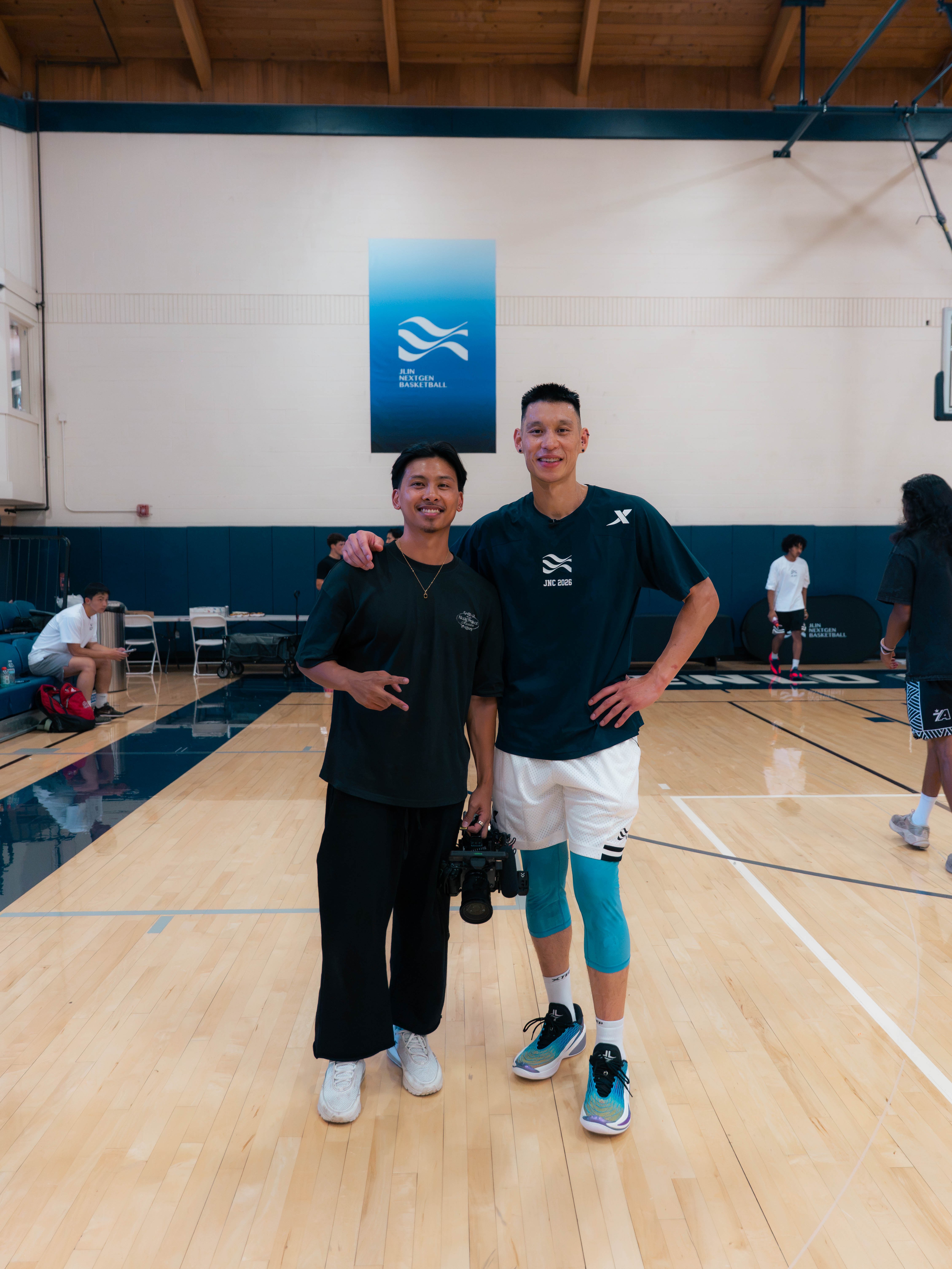 Two people standing on a basketball court, smiling, with a blue and white banner behind them, and other players in the background.