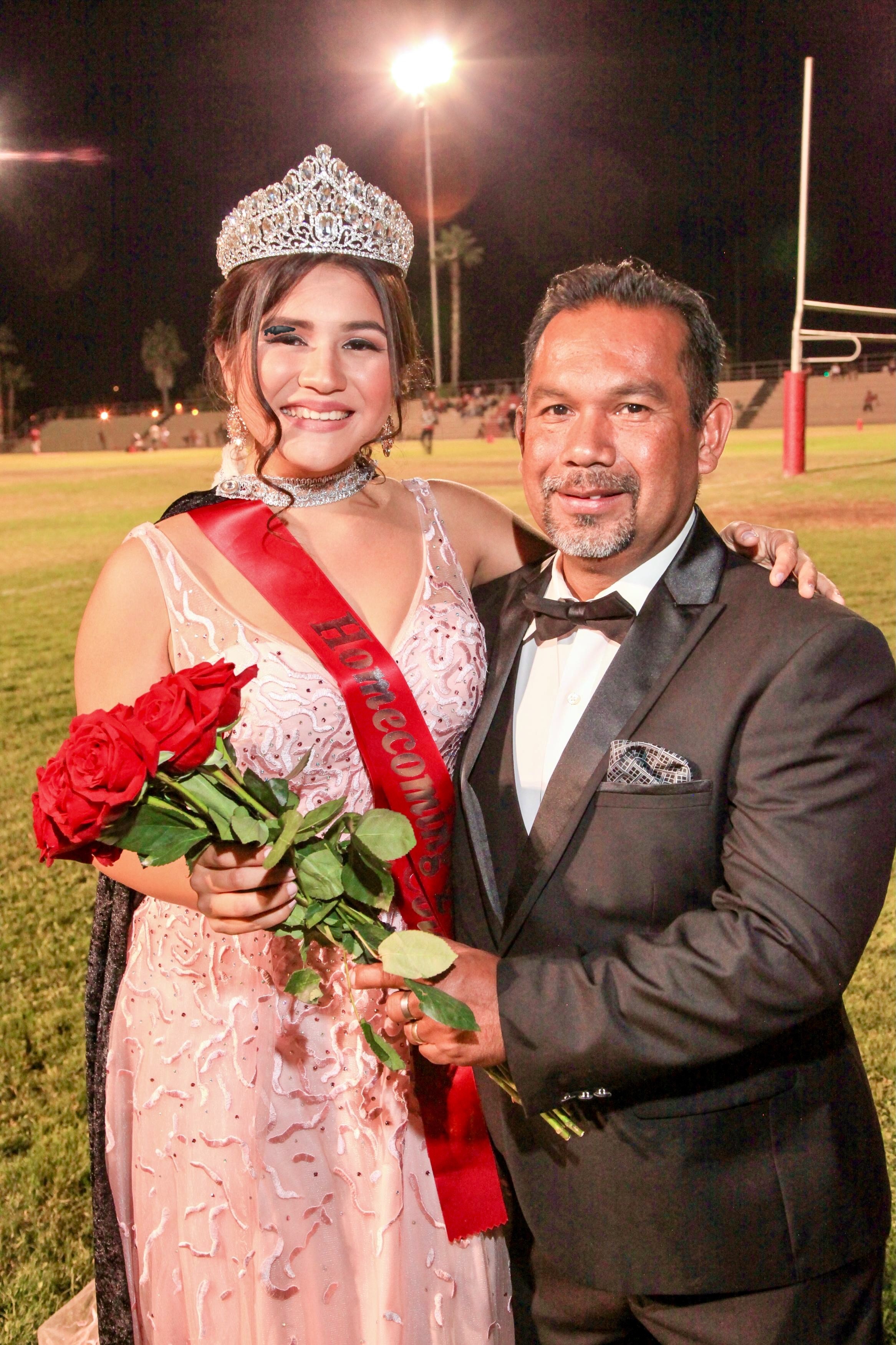 Young woman wearing a crown and sash holding red roses, standing next to a man in a tuxedo at night.