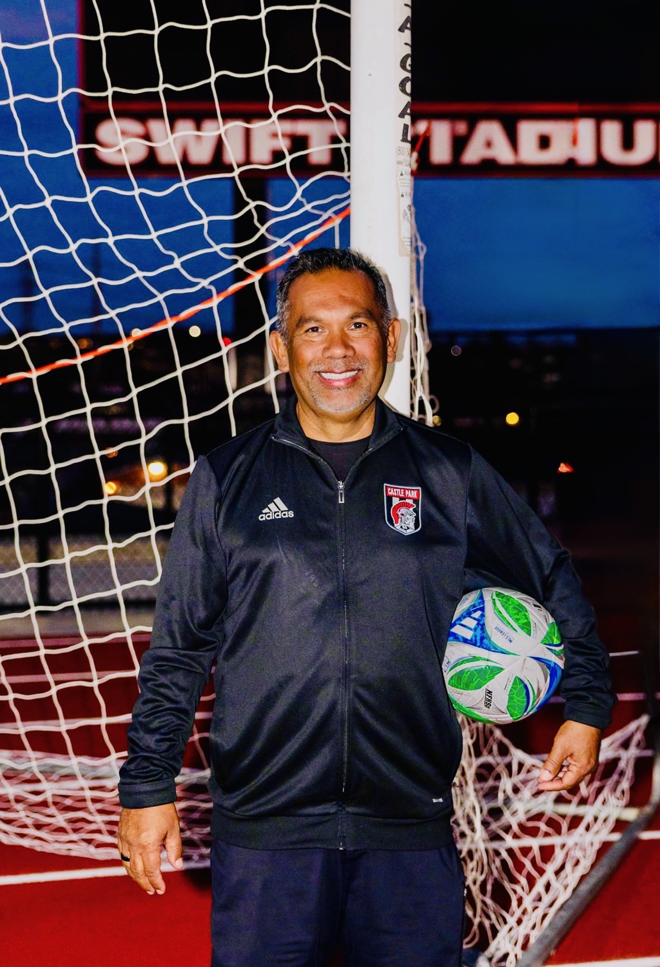Person standing in front of a soccer goal holding a ball, smiling, wearing a black jacket with a logo.