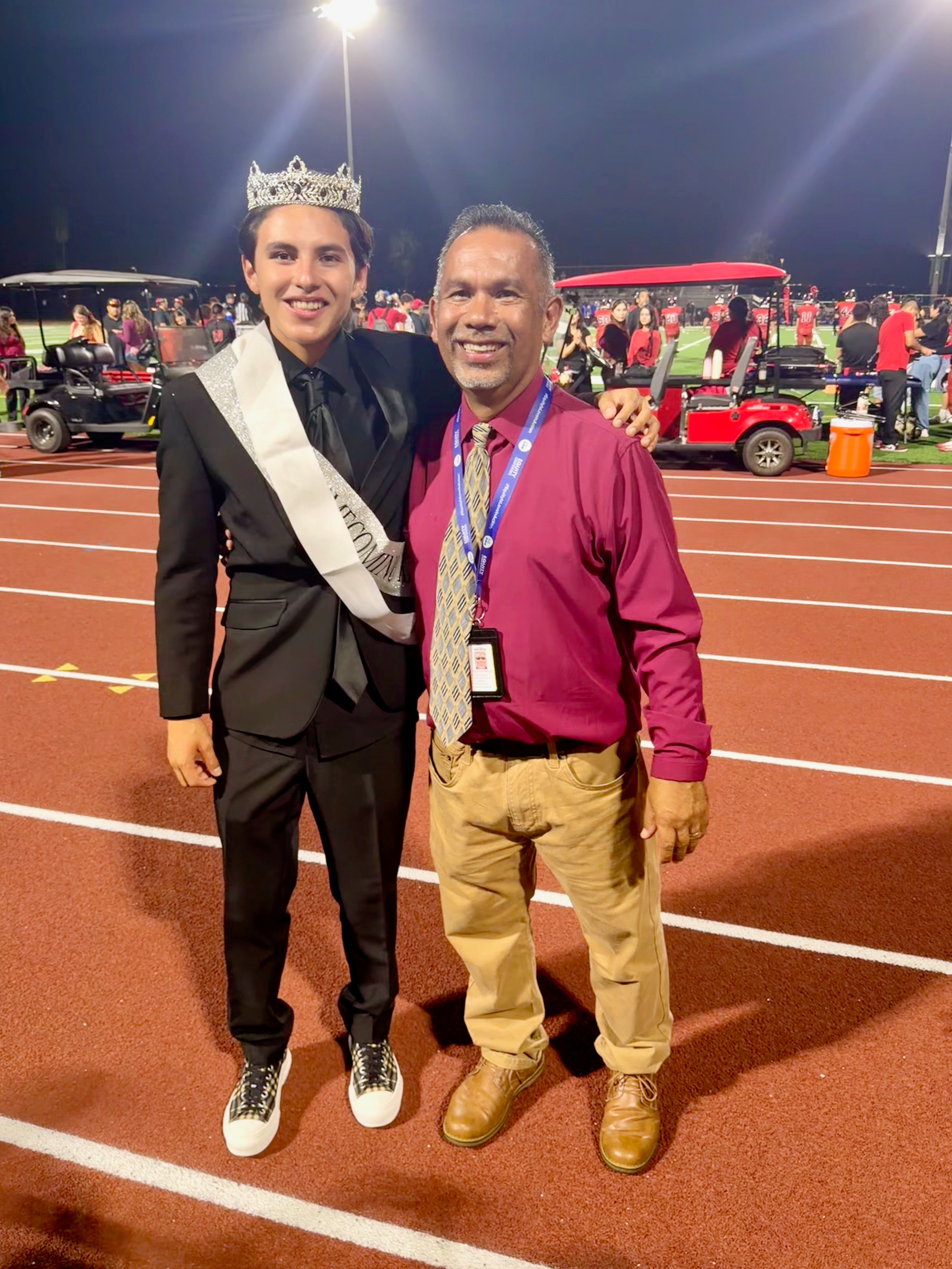 Young man wearing a crown, sash, and formal suit standing on a track with a man in a pink shirt and khakis, crowd in background.