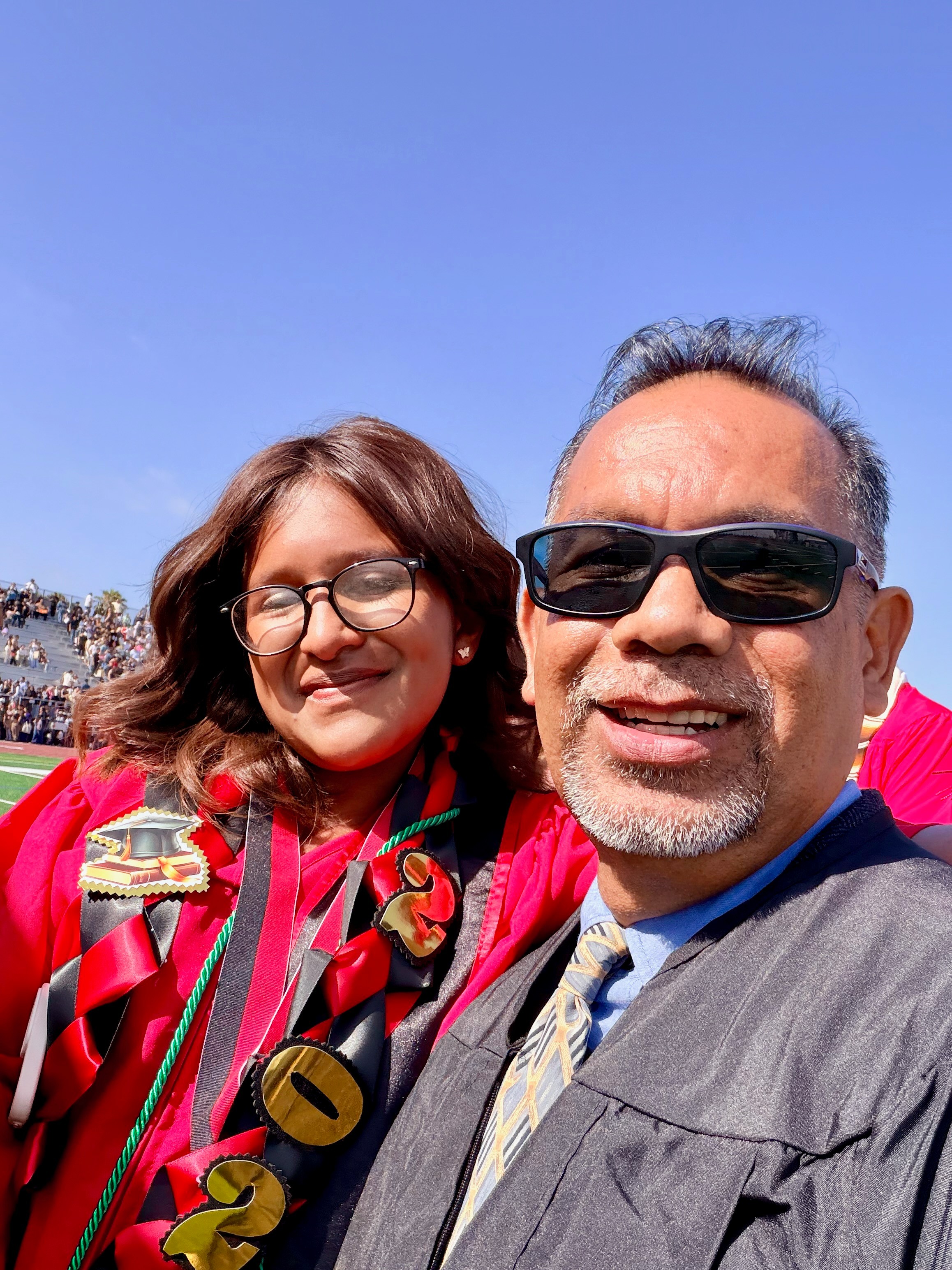 Two people smiling outdoors, one wearing glasses and a colorful striped shirt, the other wearing sunglasses and a gray robe.
