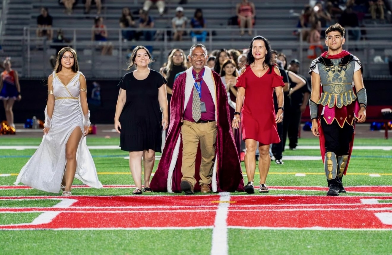 Group of five people walking on a football field, dressed in costumes, with spectators in the background.
