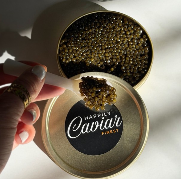 Hand holding a spoon with caviar over a round container labeled 'Happily Caviar Finest' filled with caviar.