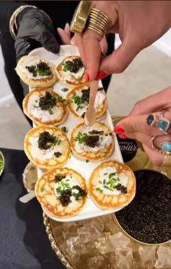 People serve small appetizers topped with black caviar and green herbs on a gold tray, with hands wearing rings and nail polish.